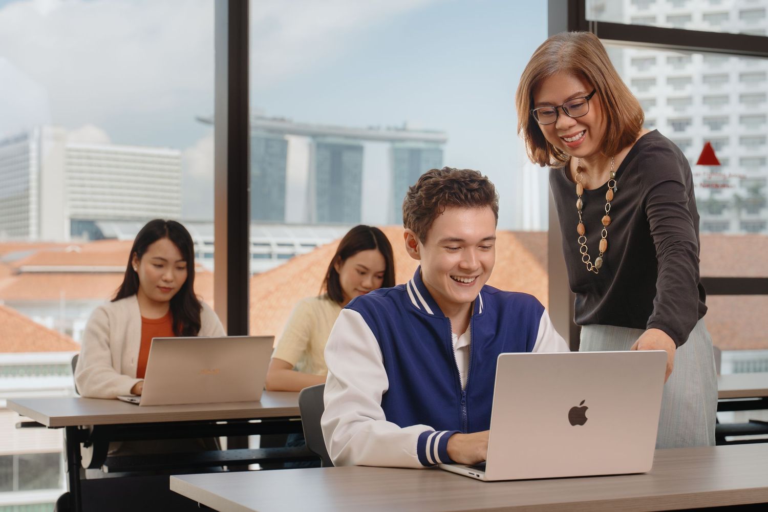 Business professionals collaborate on laptops in a modern office space with large windows.