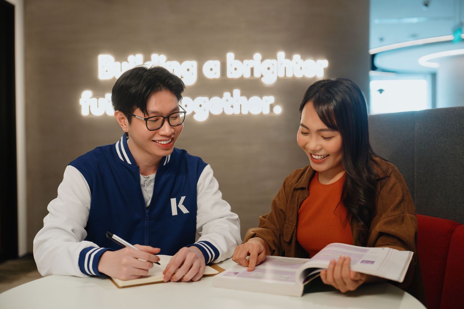 Students work together at a study table in a modern library space with bright lighting and comfortable seating.