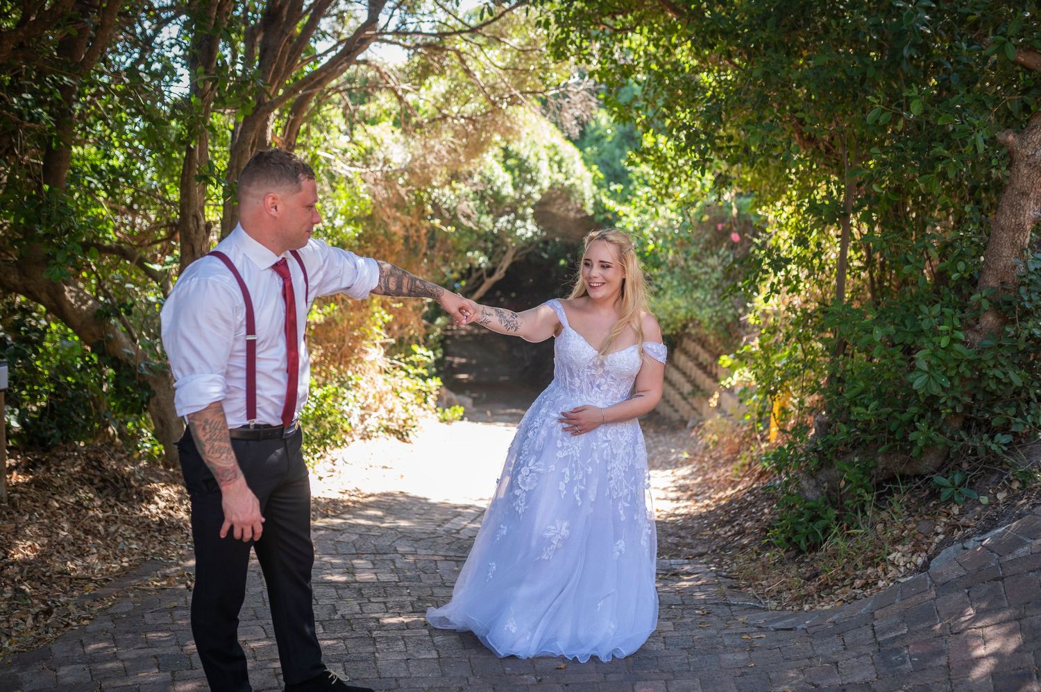 Bride in white lace gown and groom in suspenders share a romantic moment on a garden path.