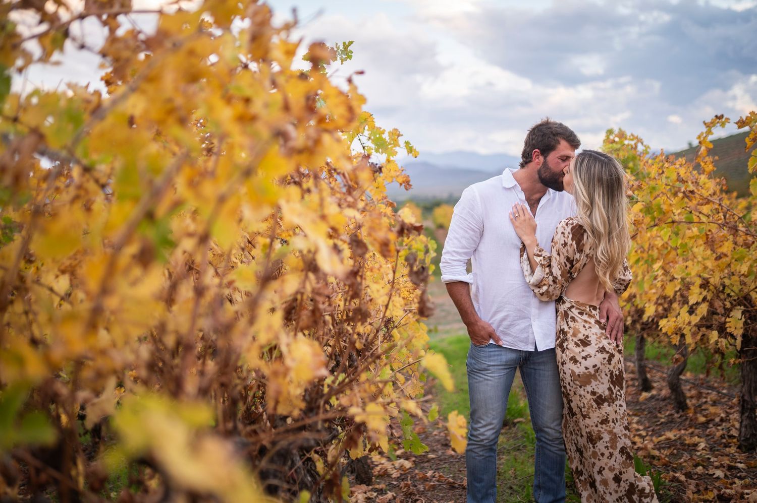 Couple kissing in autumn vineyard, surrounded by golden foliage, man in white shirt and woman in floral dress.