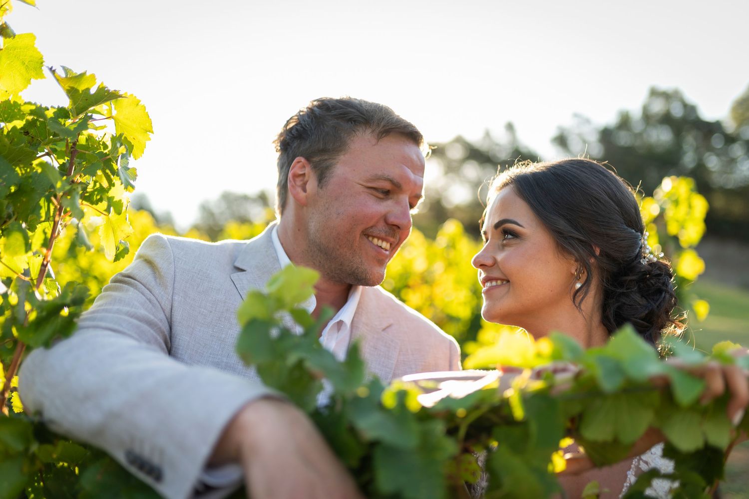 Couple smiling at each other among lush green vineyard rows in golden sunlight, perfect for a romantic winery wedding.