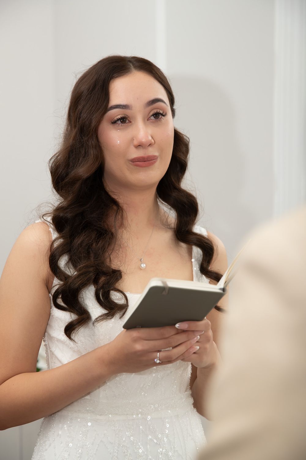 Emotional bride reads vows from a small book during an intimate indoor wedding ceremony.