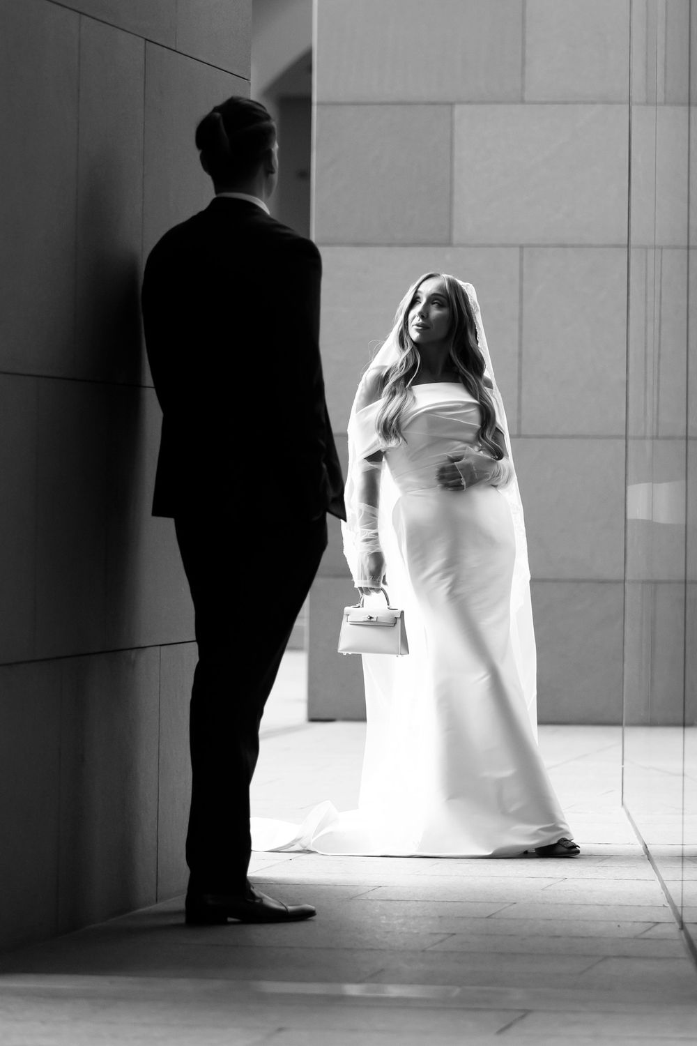 A black and white photo of a bride in a white gown and veil standing near her suited groom.