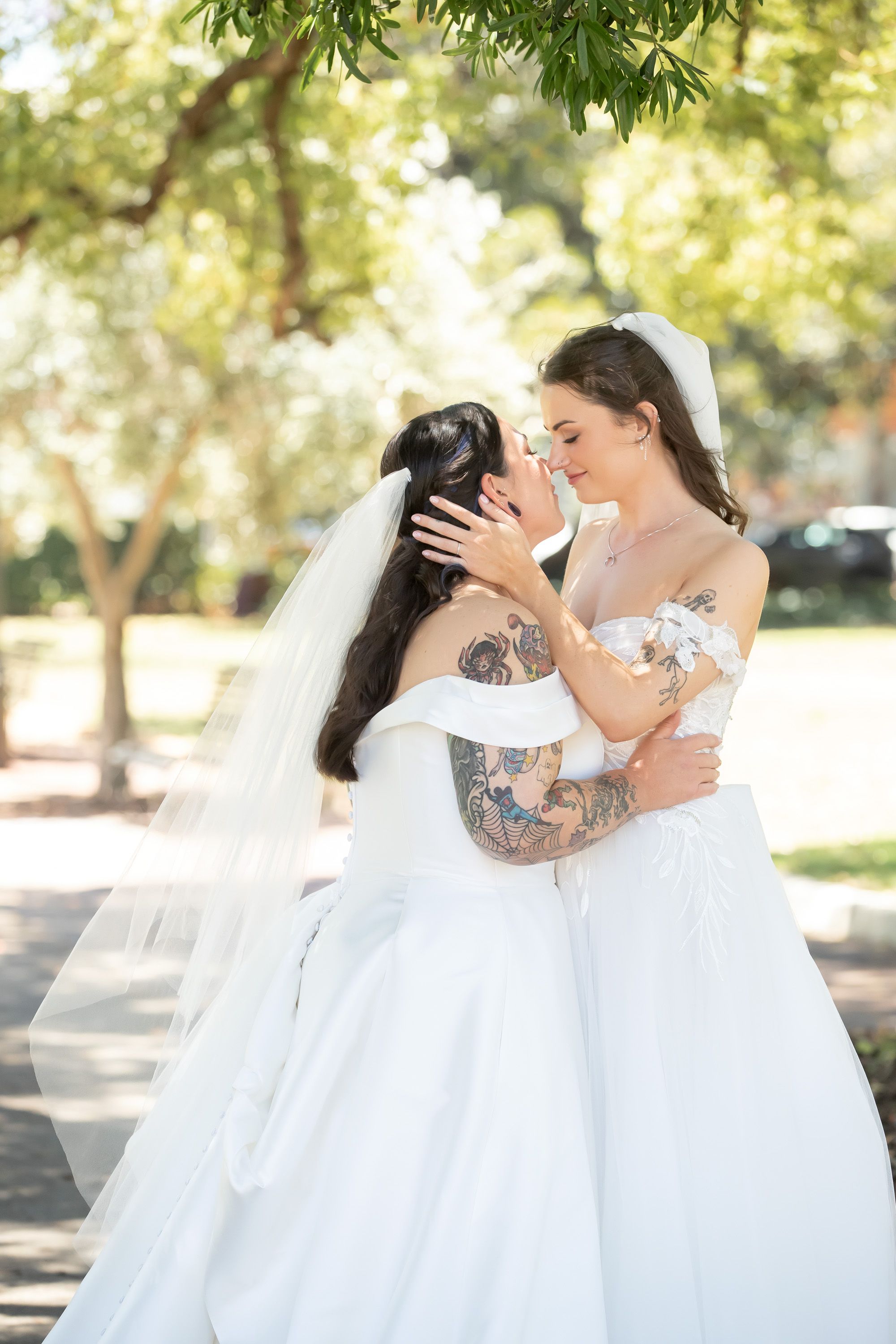 Two brides in white gowns and veils embrace tenderly outdoors under sunlit trees.