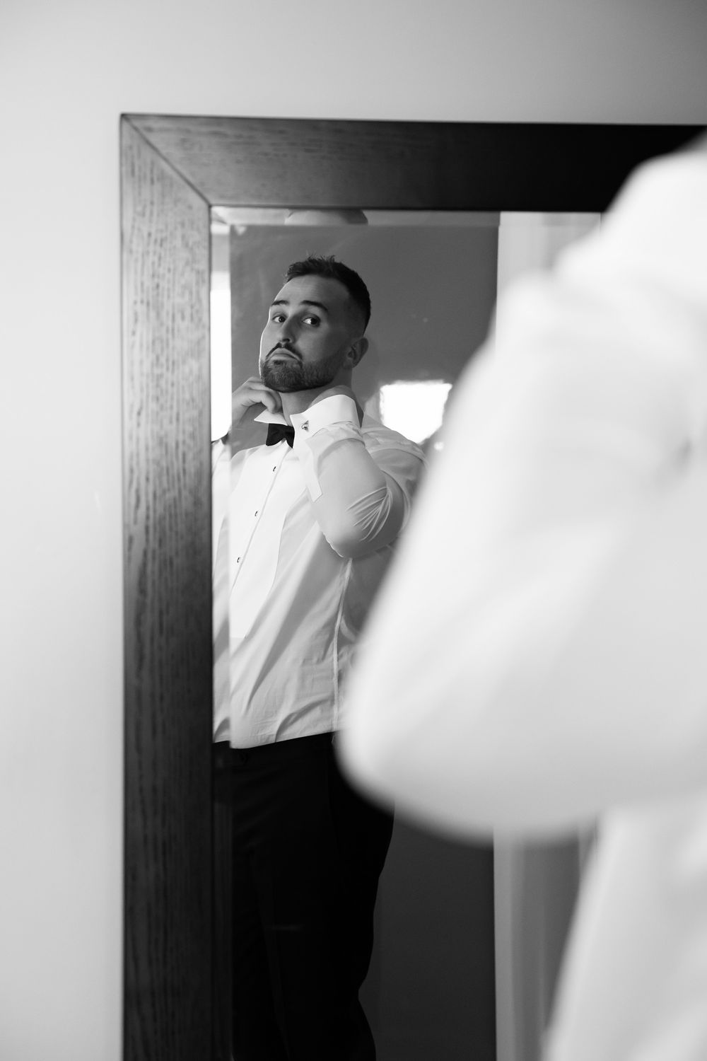 Groom adjusts bow tie while checking reflection in mirror, black and white wedding photo.