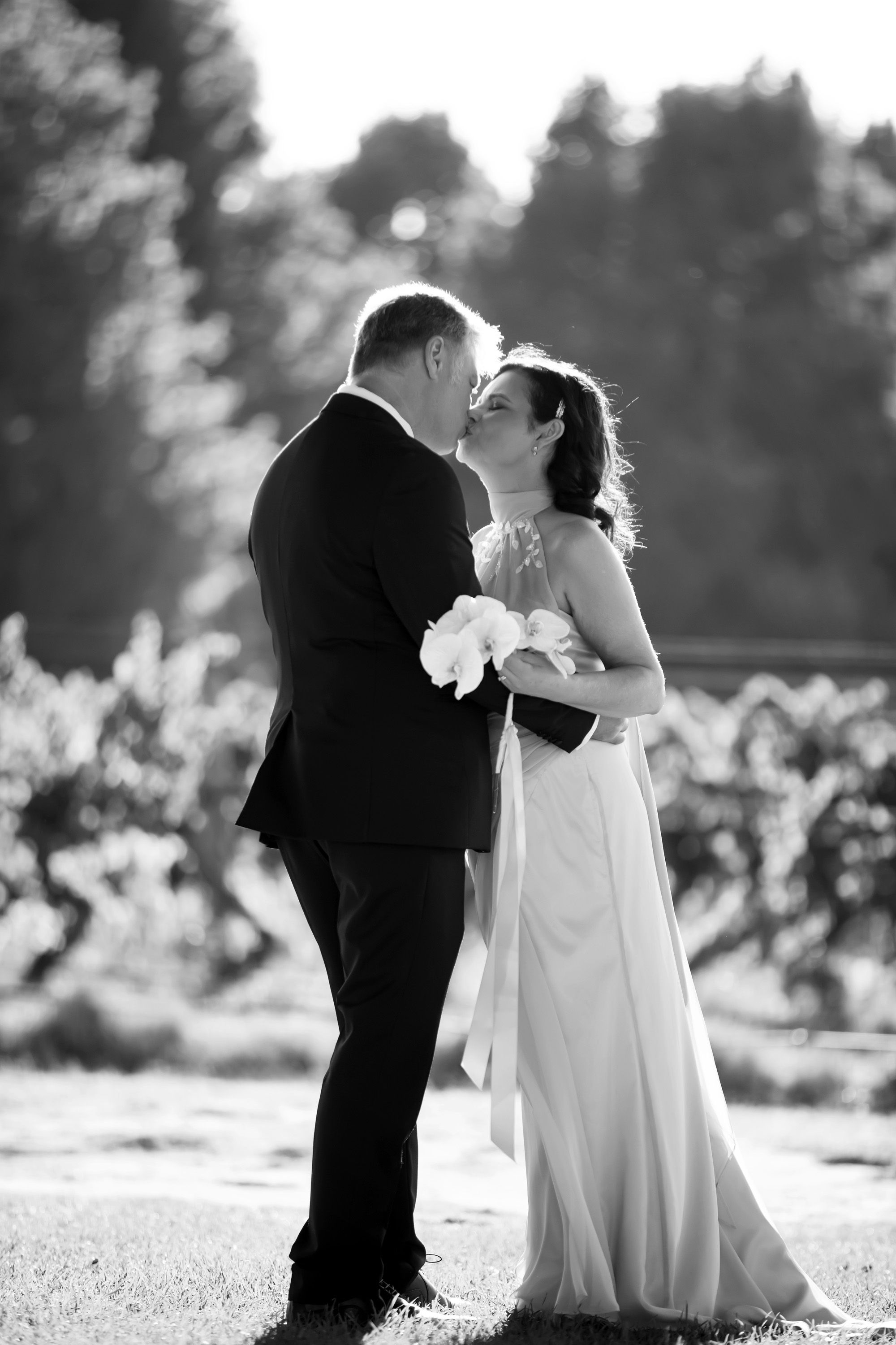 Bride and groom share a kiss outdoors in black and white wedding portrait with soft bokeh.