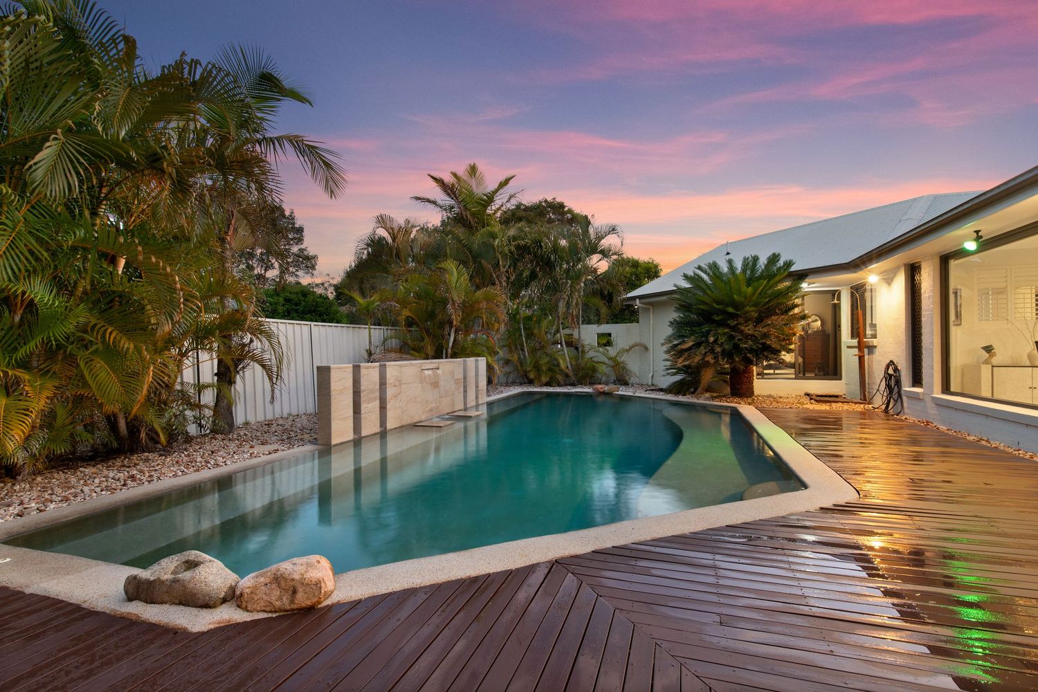 Sparkling pool with timber deck and tropical palms beside modern home at dusk.