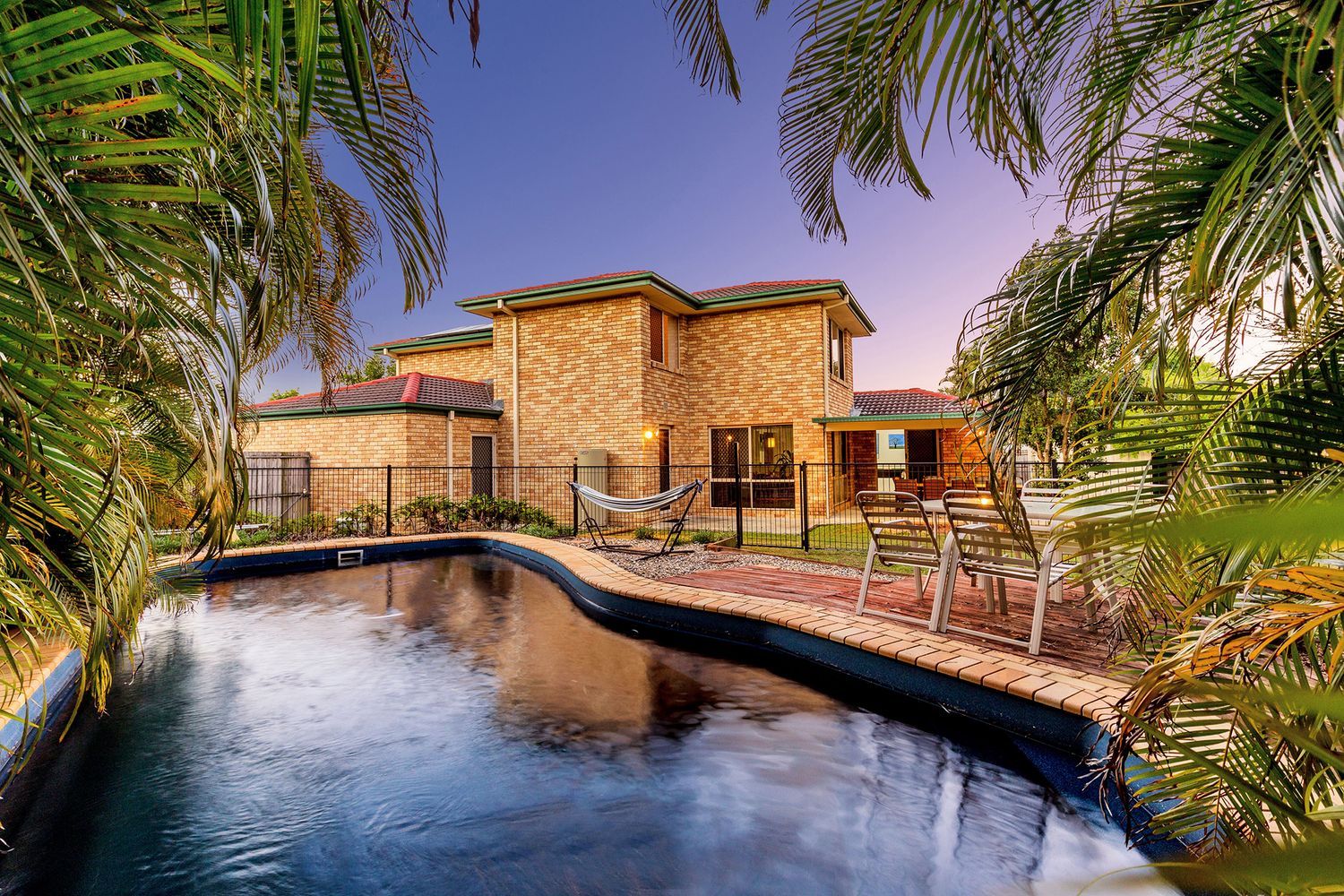 Lagoon-style pool with brick paving, palm trees, and two-story brick home at twilight.