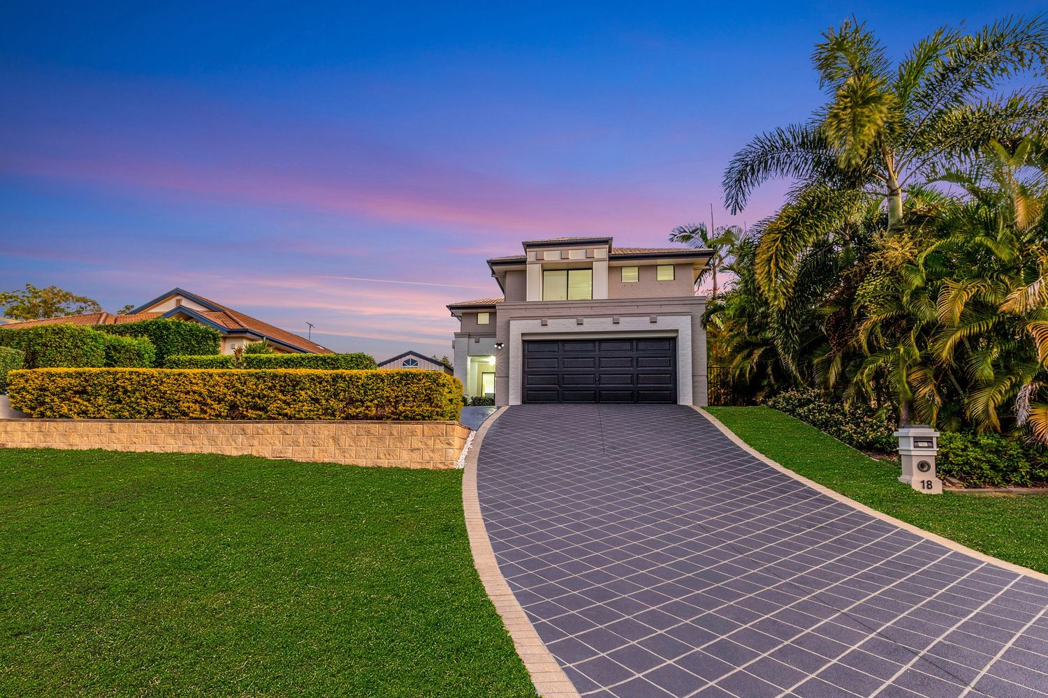 Modern two-story home with dark garage door, paved driveway, lush lawn, and palm trees at dusk.