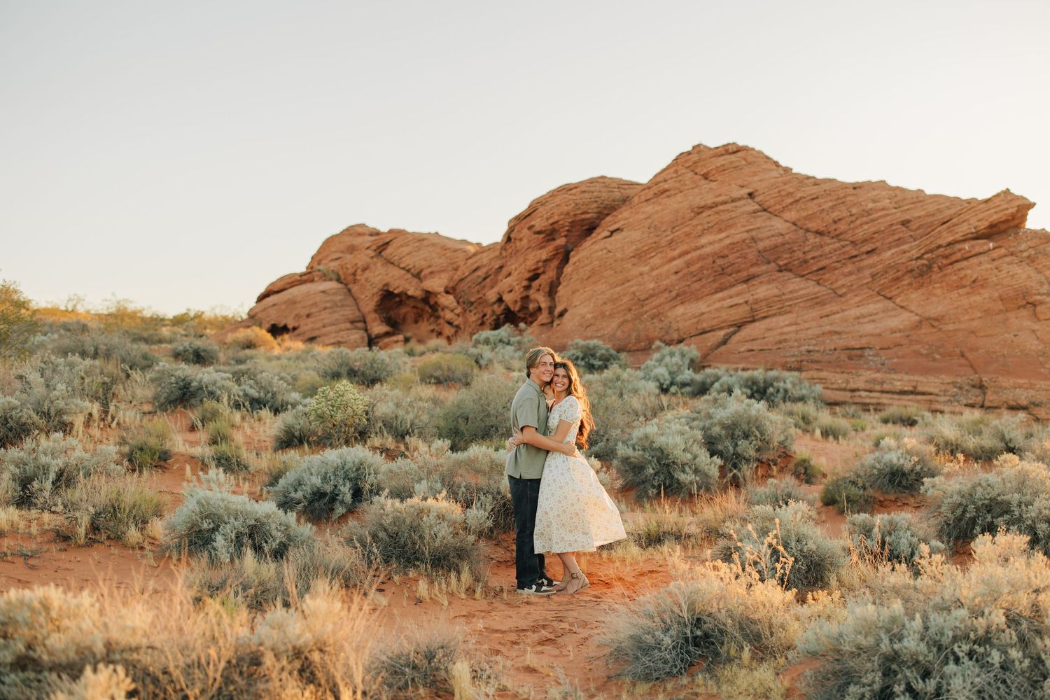 Couple in western attire embracing against stunning red rock formations in a desert landscape at sunset.