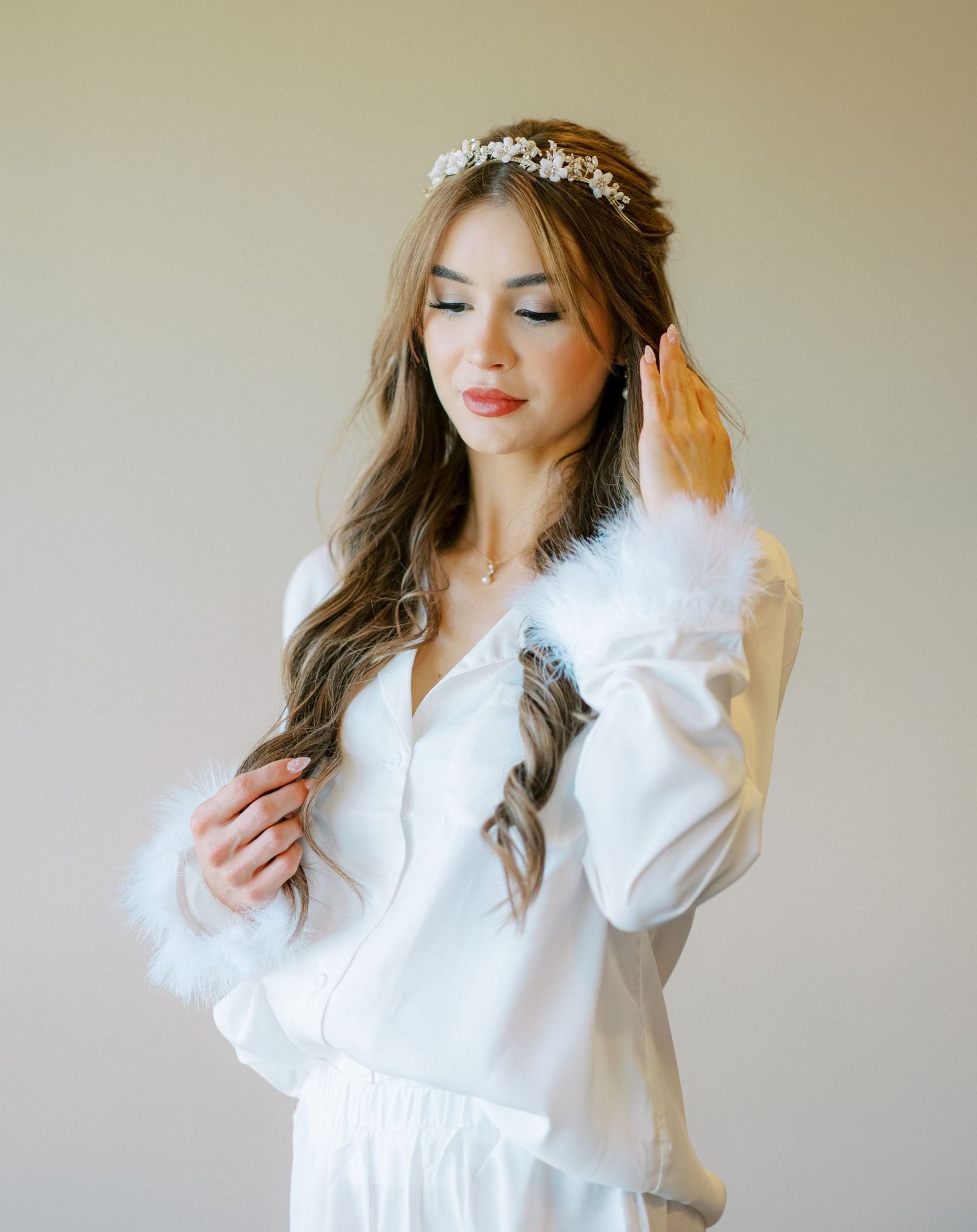 Bridal portrait in white feather-trimmed dress and crystal headpiece against neutral background.