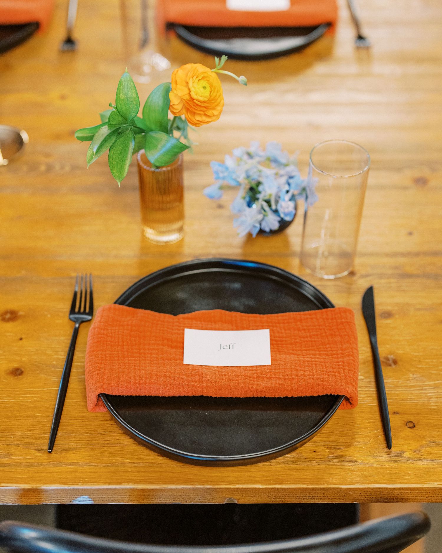 Modern place setting with orange napkin and black plate on wooden table with floral accents.