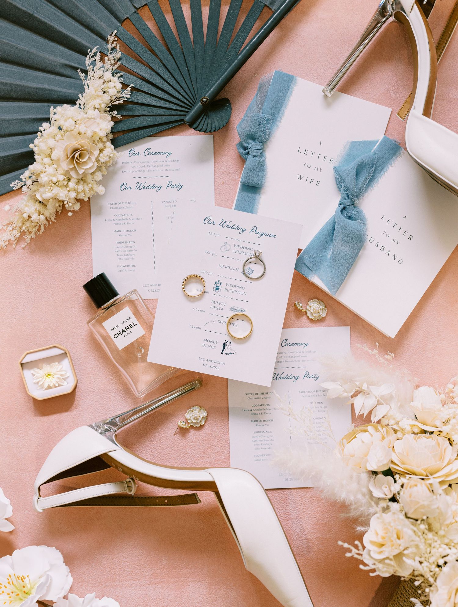 Wedding details flatlay with invitations, perfume, rings, and shoes on a pink background.