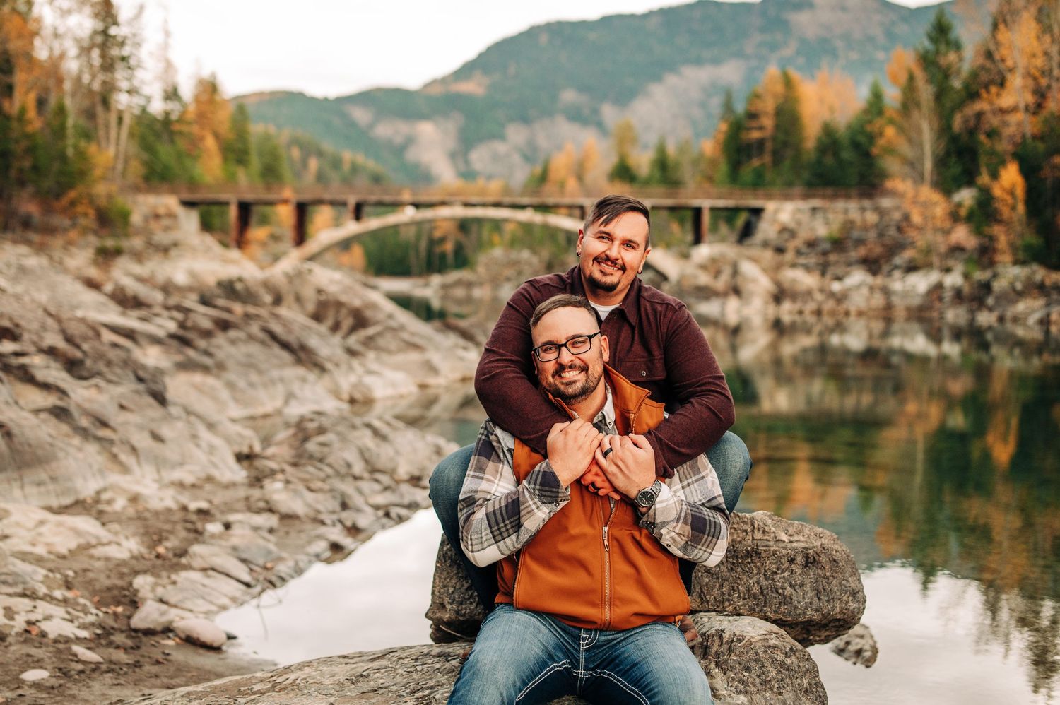 Two people share a joyful moment while sitting on rocks by a mountain lake with an arched bridge in the background.