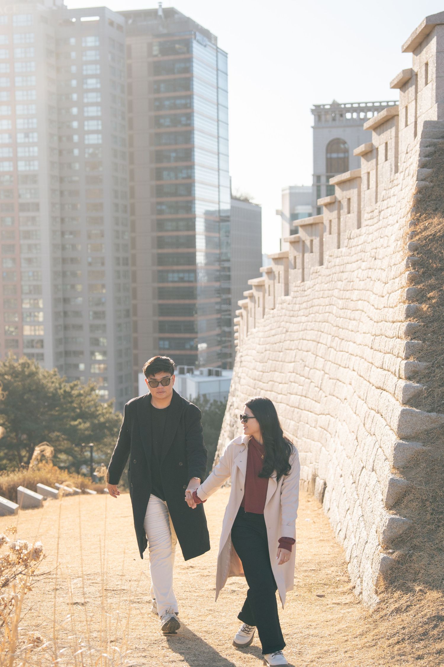 A couple walks hand in hand along a sunlit stone wall path with high-rise buildings visible in the background.