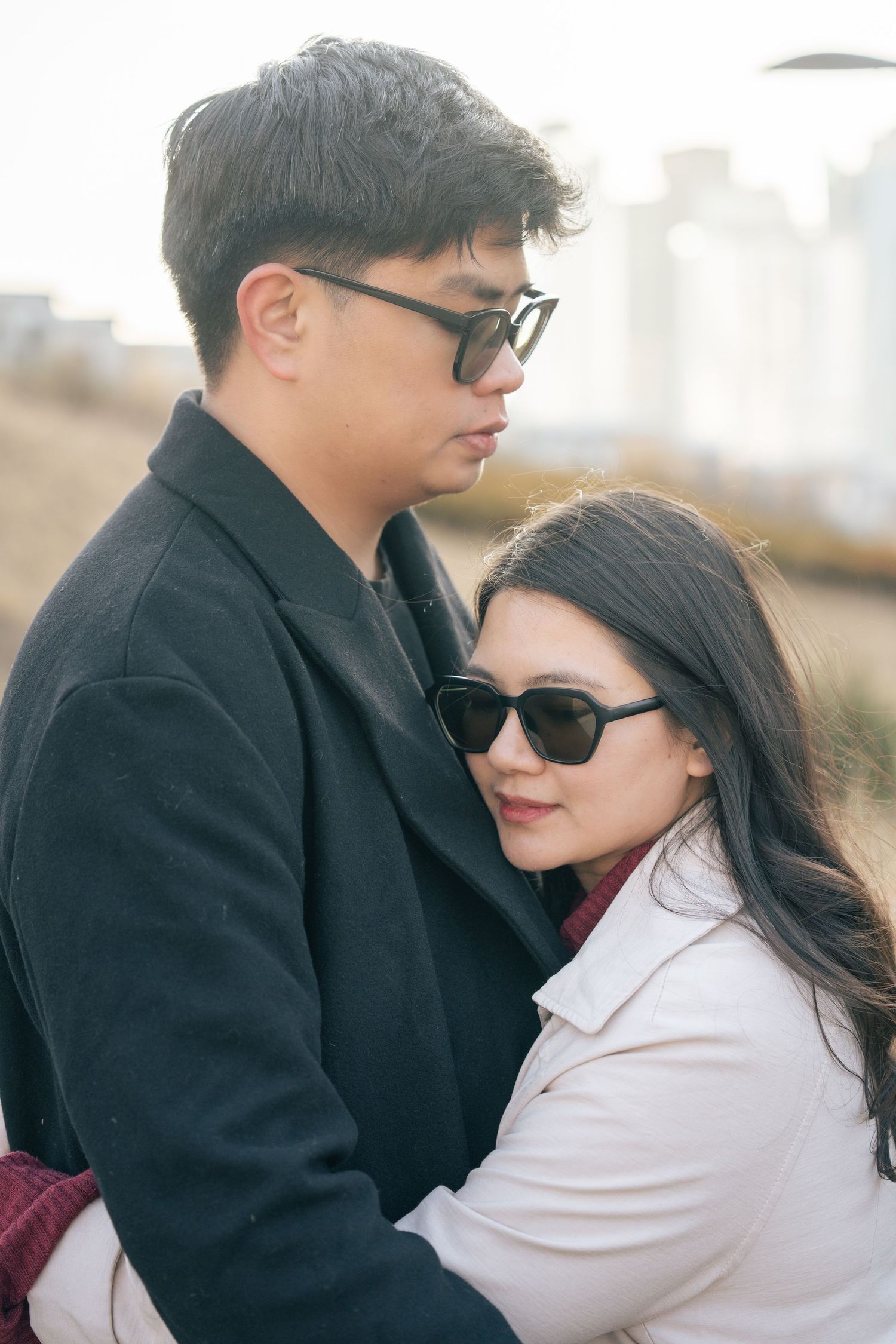 A couple in dark coats and sunglasses embrace outdoors in a romantic moment.