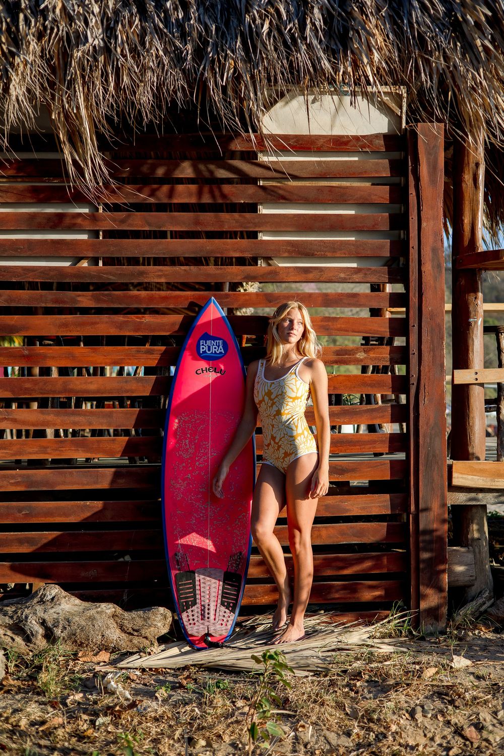 A pink surfboard leans against a wooden fence wall at sunset while someone stands nearby in a swimsuit.