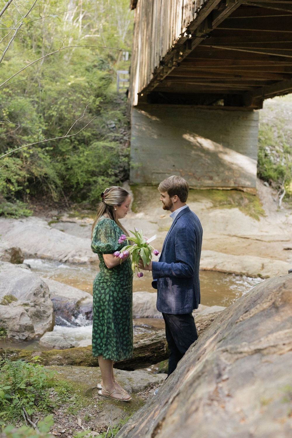 Couple standing on rocks by a creek beneath a wooden bridge, woman in green dress holding flowers.
