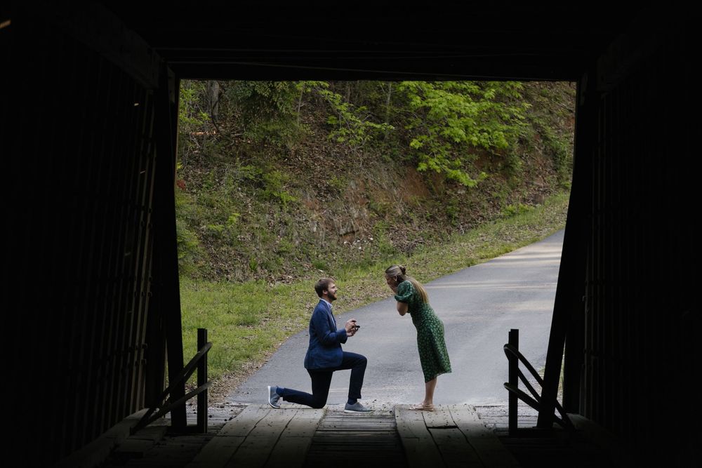 Man proposing on one knee to woman in green dress at covered bridge entrance surrounded by lush forest.