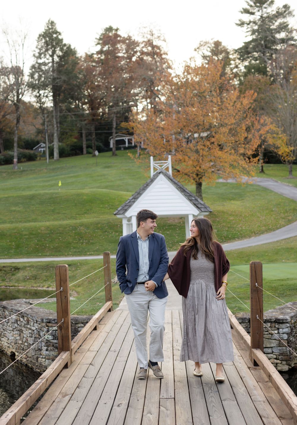 Couple walking together on wooden bridge with white gazebo and fall foliage in background.