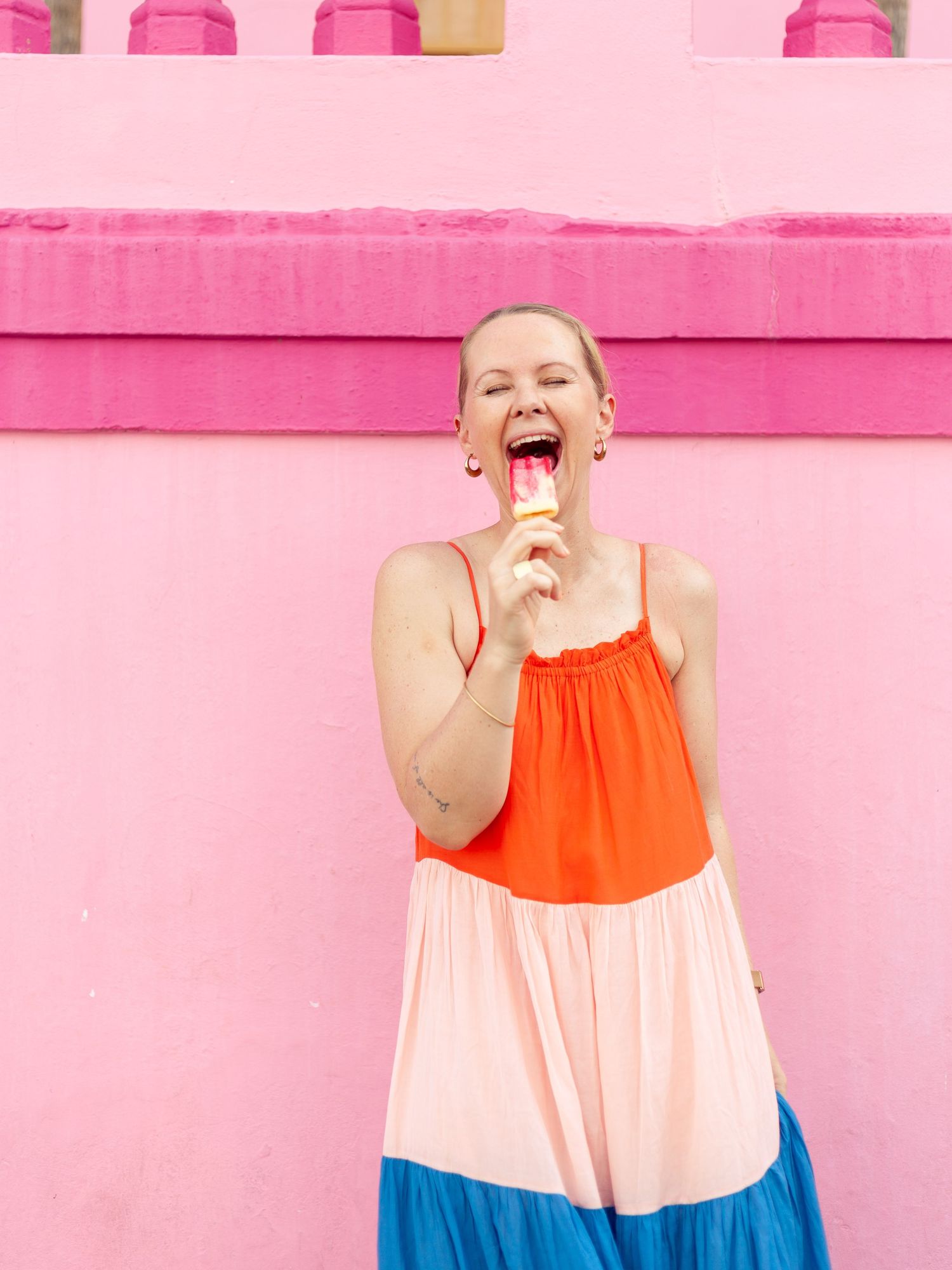 Person enjoying ice cream against pink wall background in colorful dress.