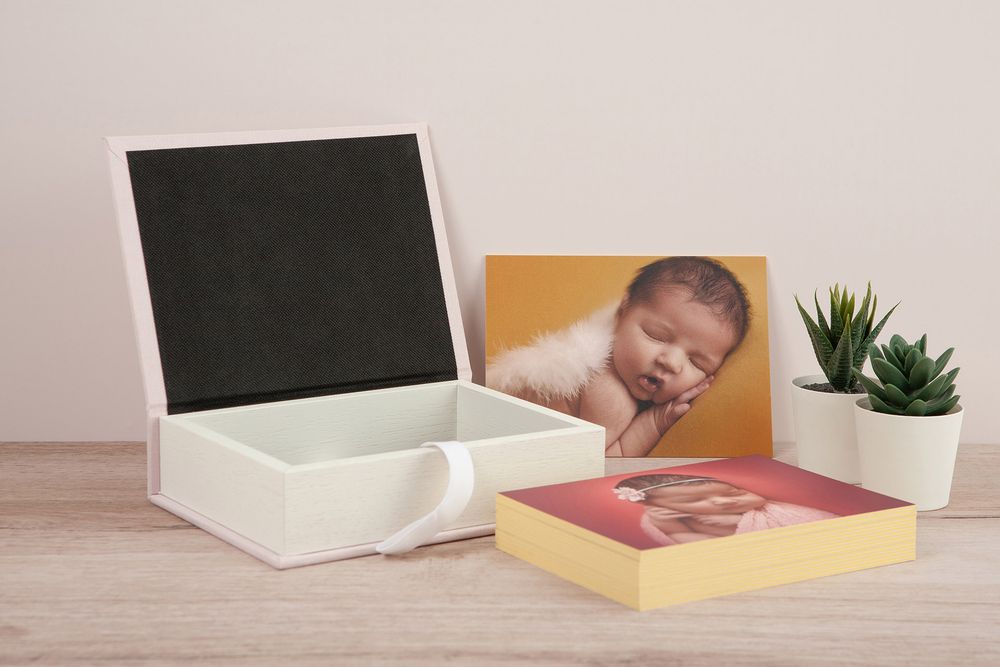 Hands opening a white box with a photo album and small potted plant on wooden surface.
