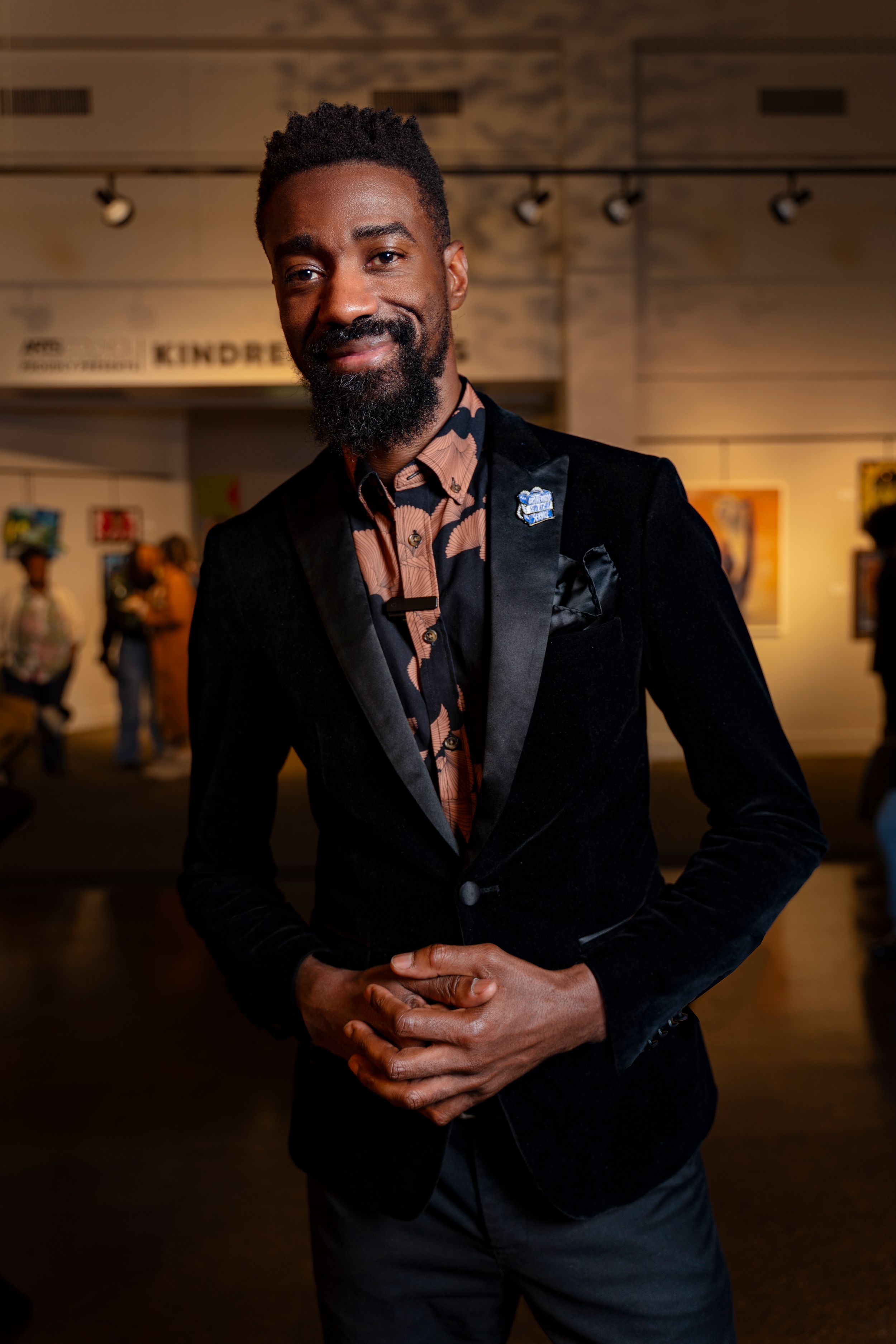 A person wearing a black suit and tie poses in an art gallery space with dramatic lighting.