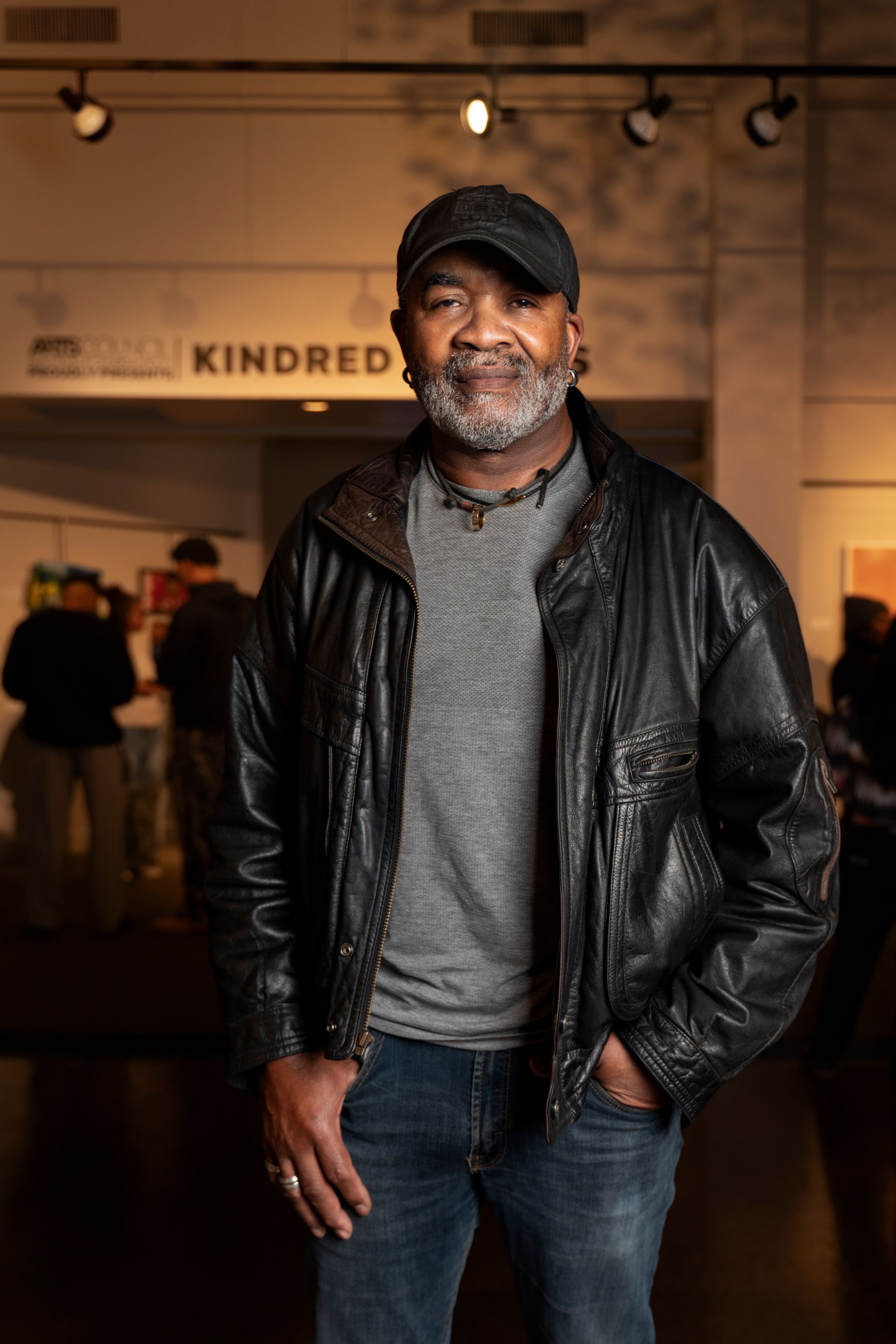 A person wearing a black leather jacket and gray shirt stands in an art gallery space with track lighting overhead.