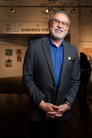 Person in dark suit jacket and blue shirt poses in front of gallery wall with spotlights.