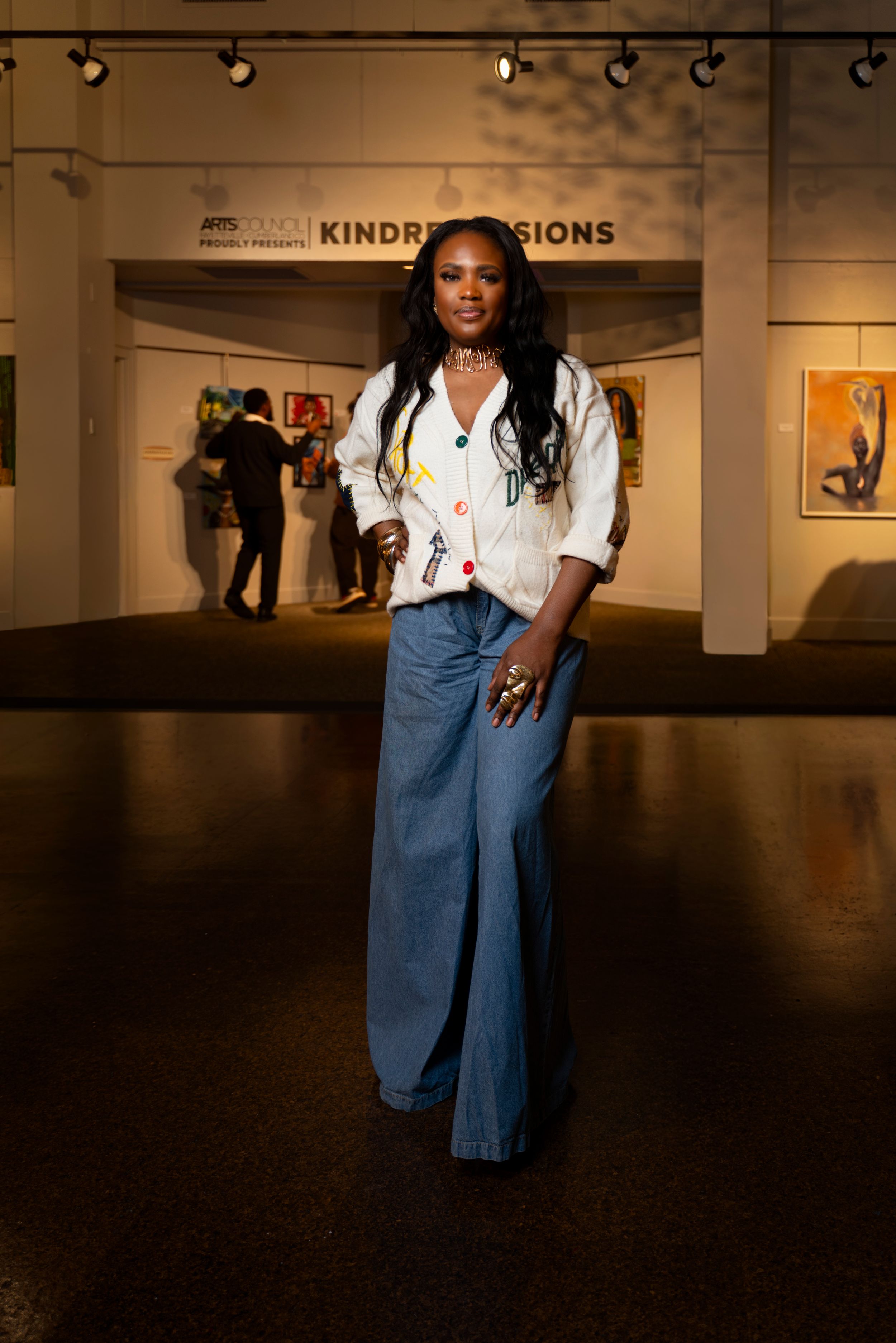 Person in denim overalls and white t-shirt stands in art gallery space with dark floors and white walls.