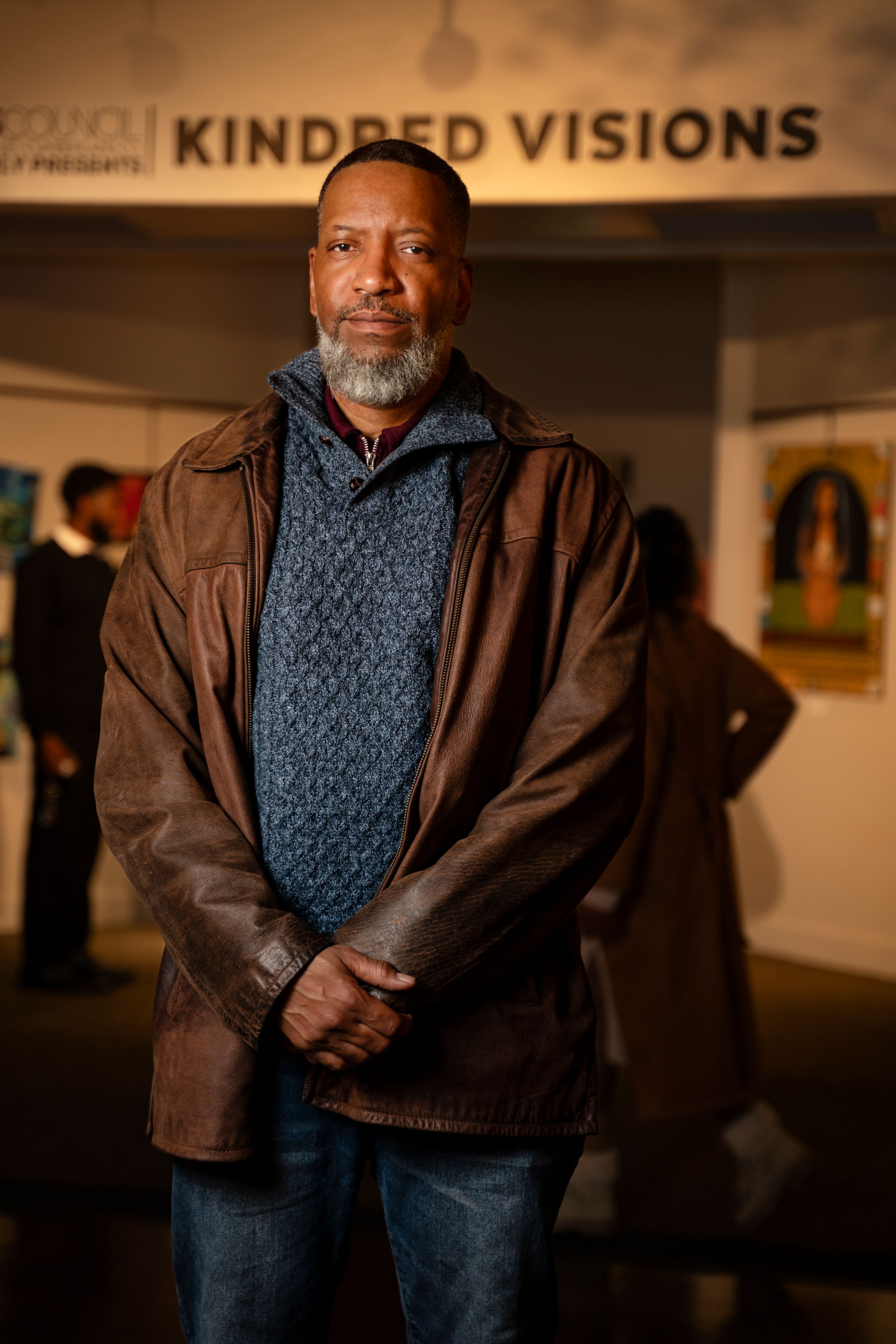 Person wearing a brown jacket and knit sweater stands in an art gallery with 'Kindred Visions' signage visible.