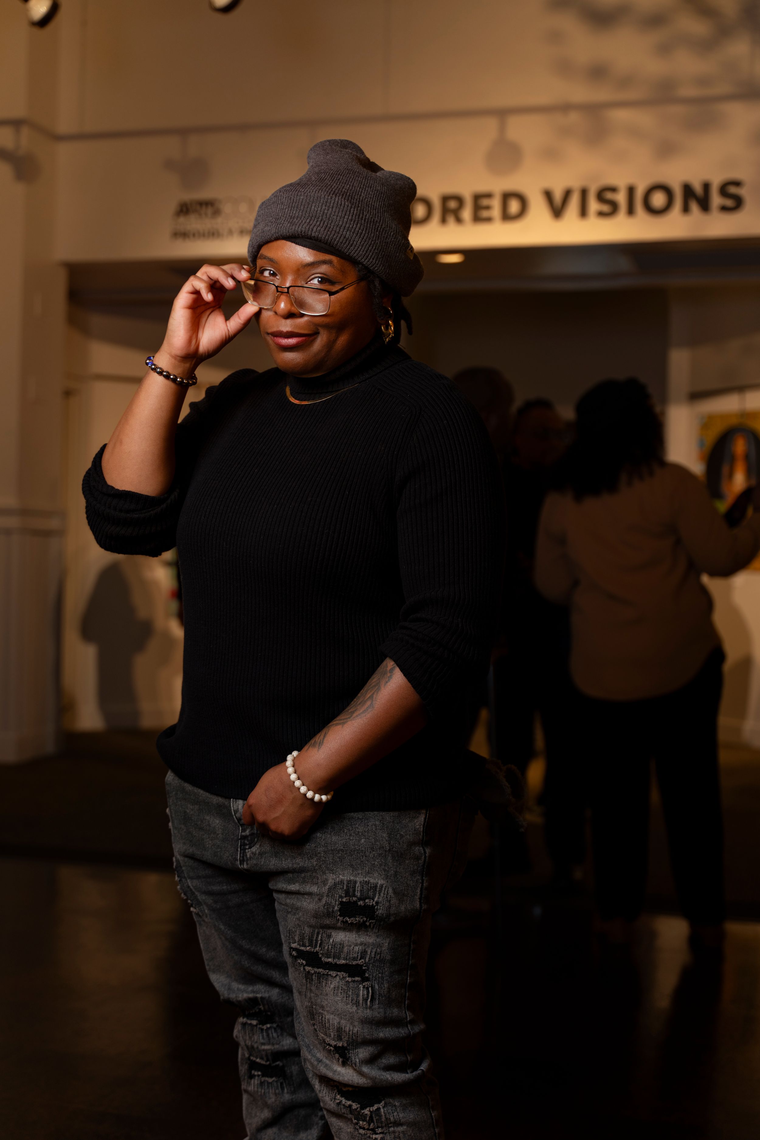 Person wearing a black beanie, glasses and sweater poses casually against an indoor wall with blurred signage.