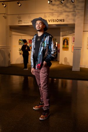 A stylishly dressed person in a hat and shiny jacket poses in an art gallery exhibition space.
