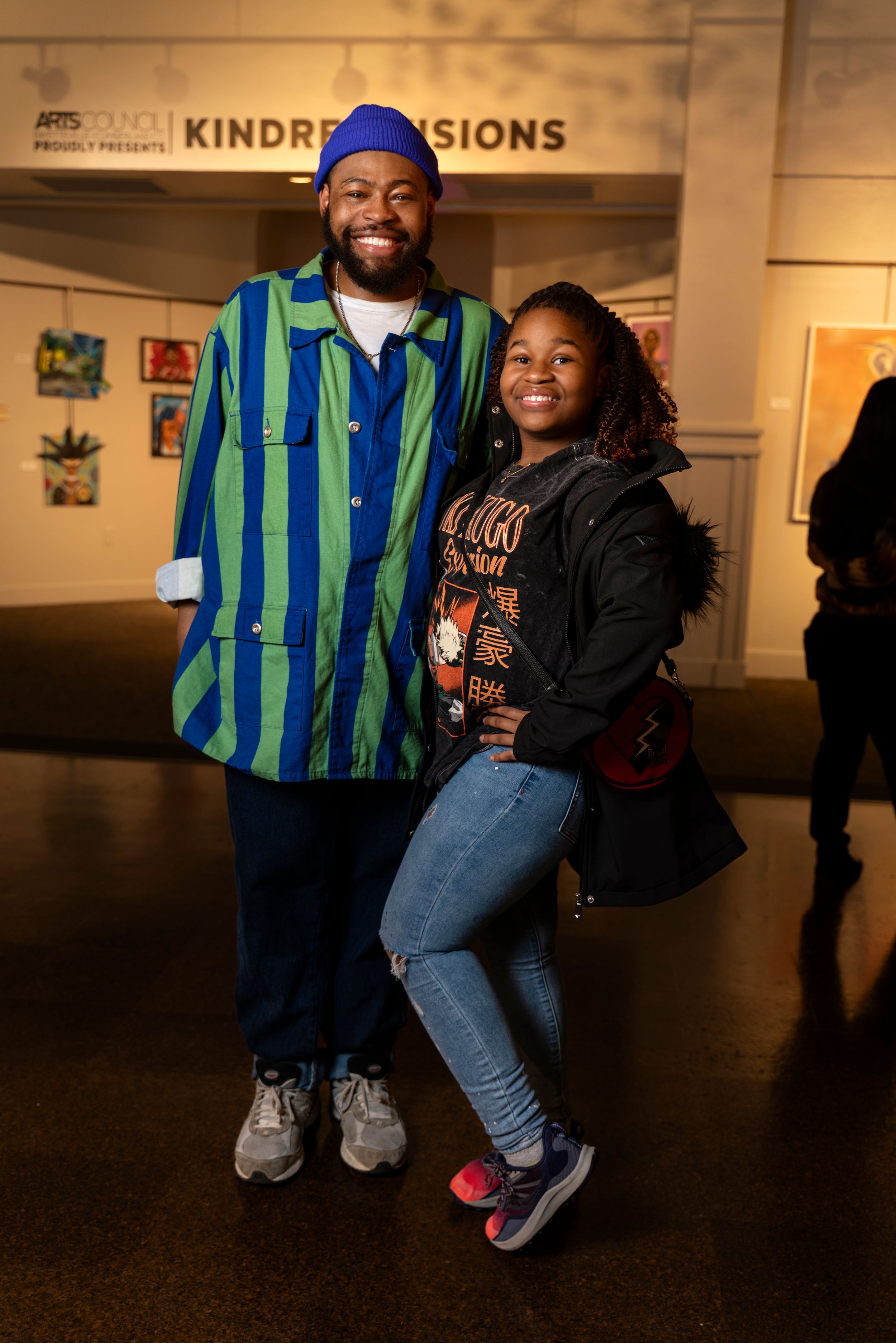 Two people pose together in front of 'Kindred Visions' gallery sign, one wearing striped shirt and jeans.