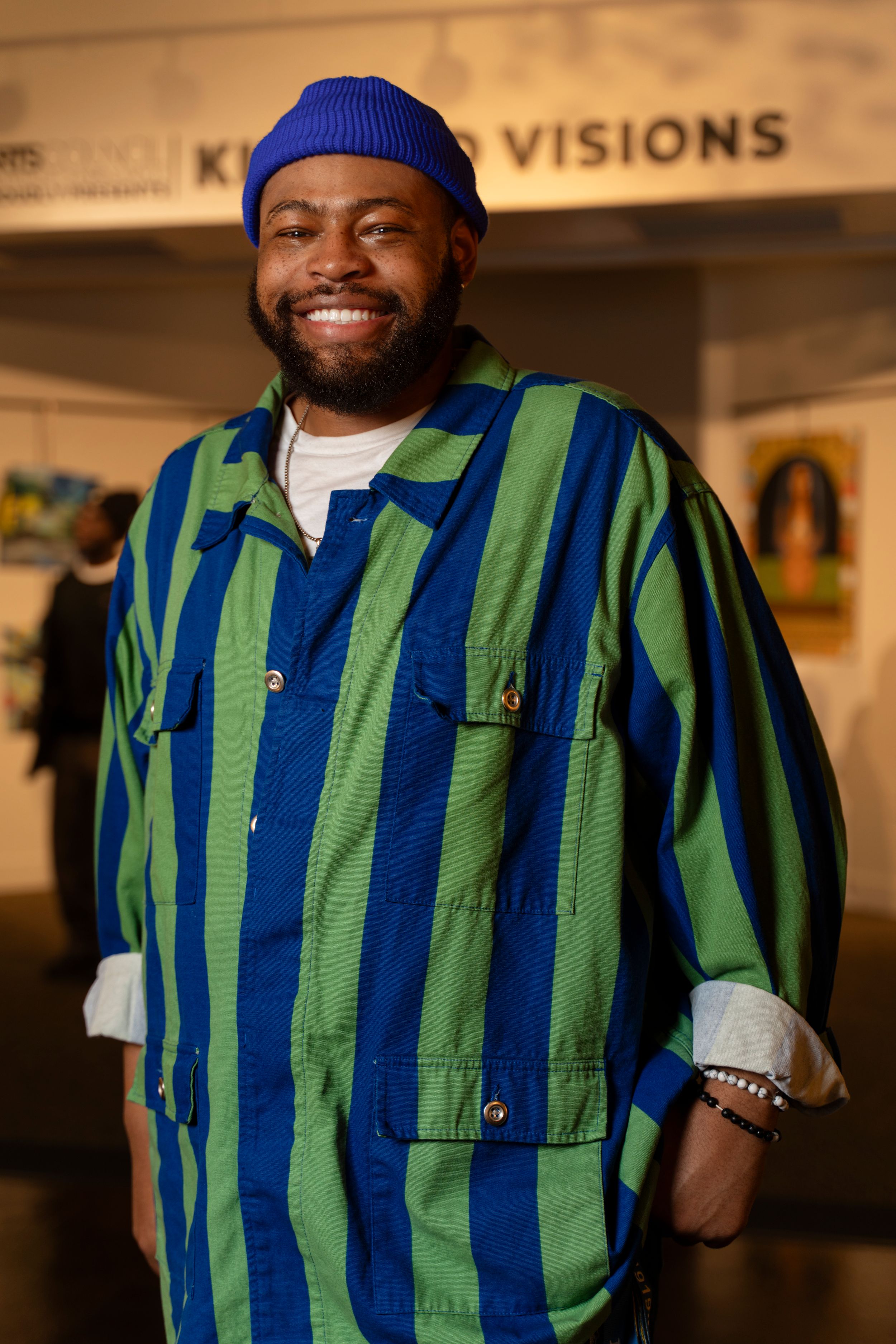 A person in a blue beanie and striped green and blue vest smiles warmly in an indoor gallery setting.