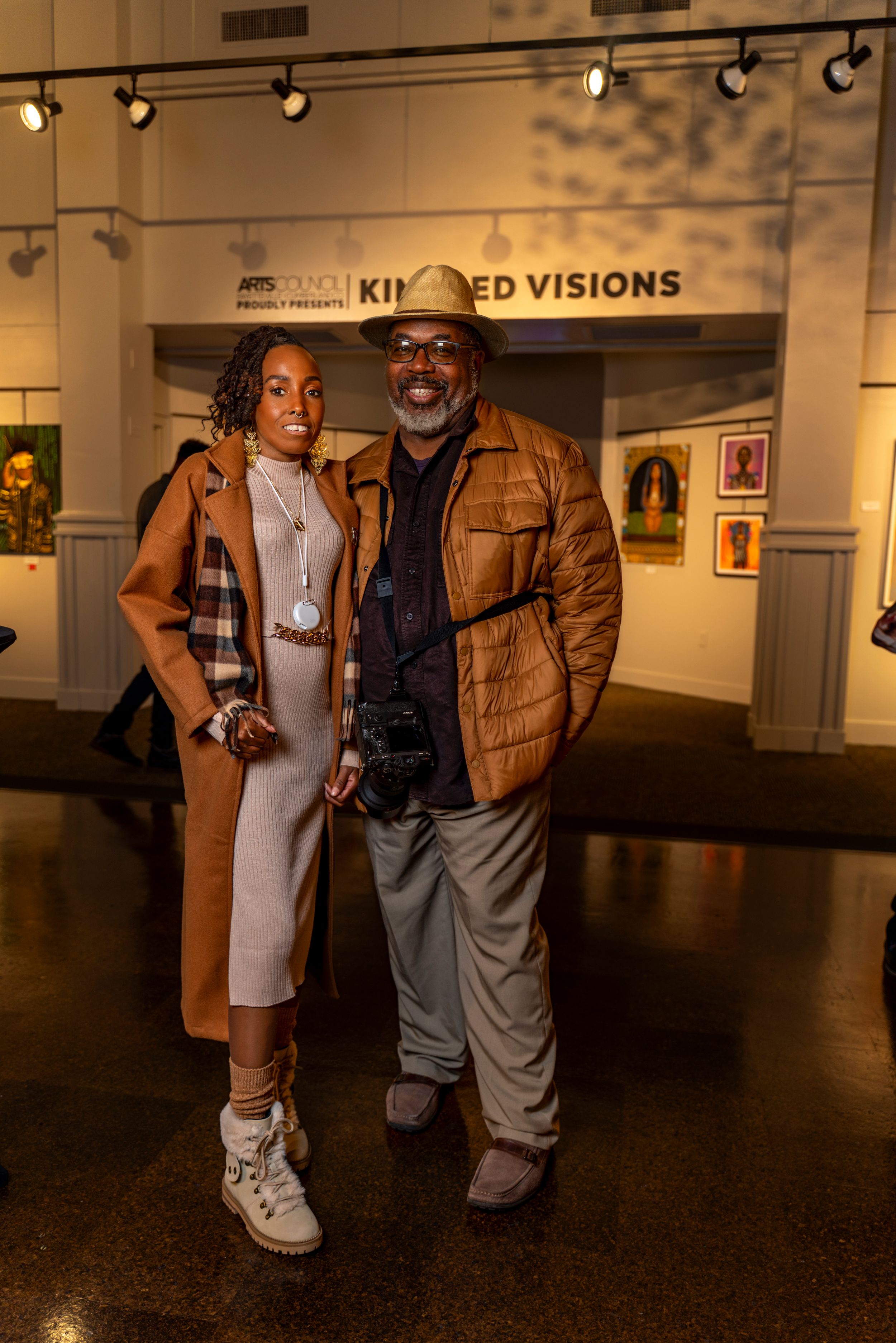 Two people stand together in an art gallery space with track lighting and white walls displaying artwork.