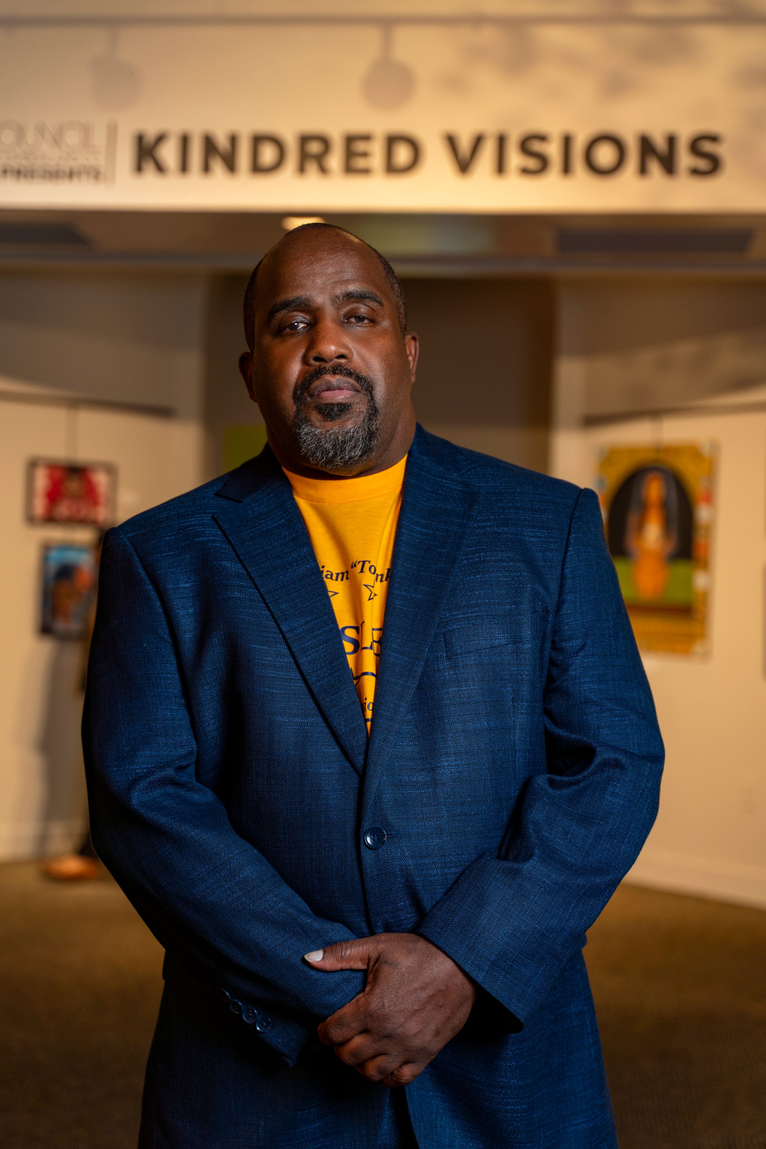 A person in a navy blue blazer and yellow shirt stands in an art gallery under the Kindred Visions sign.