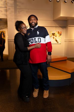 Two people embrace while kneeling in a dimly lit art gallery space with dark floors.