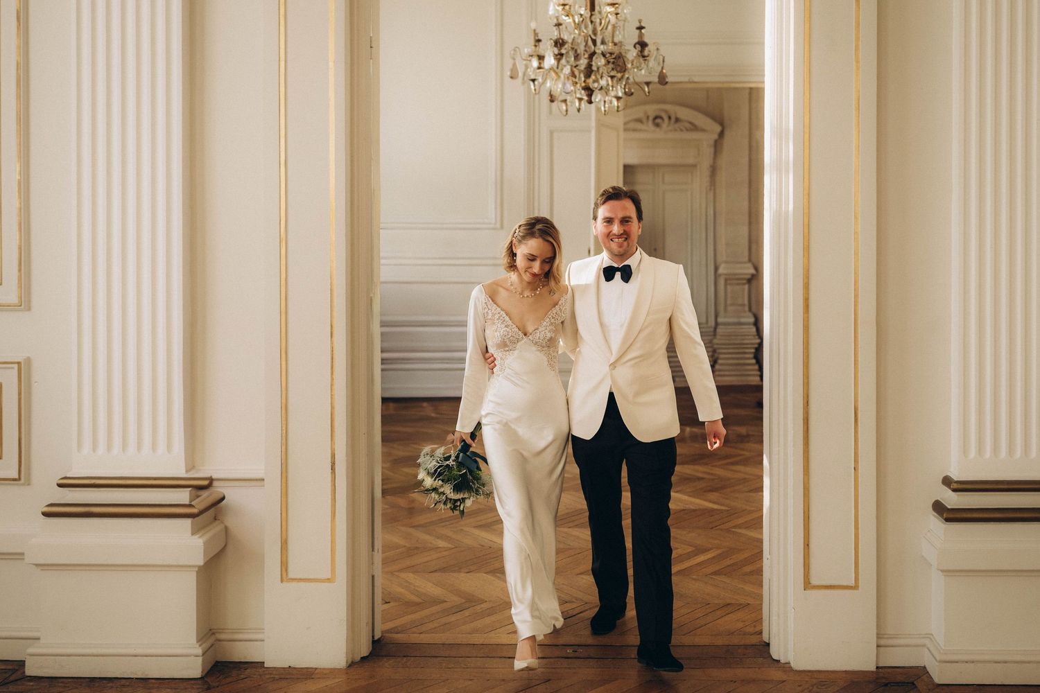 Elegant wedding couple poses in a grand white ballroom with ornate columns and chandeliers.