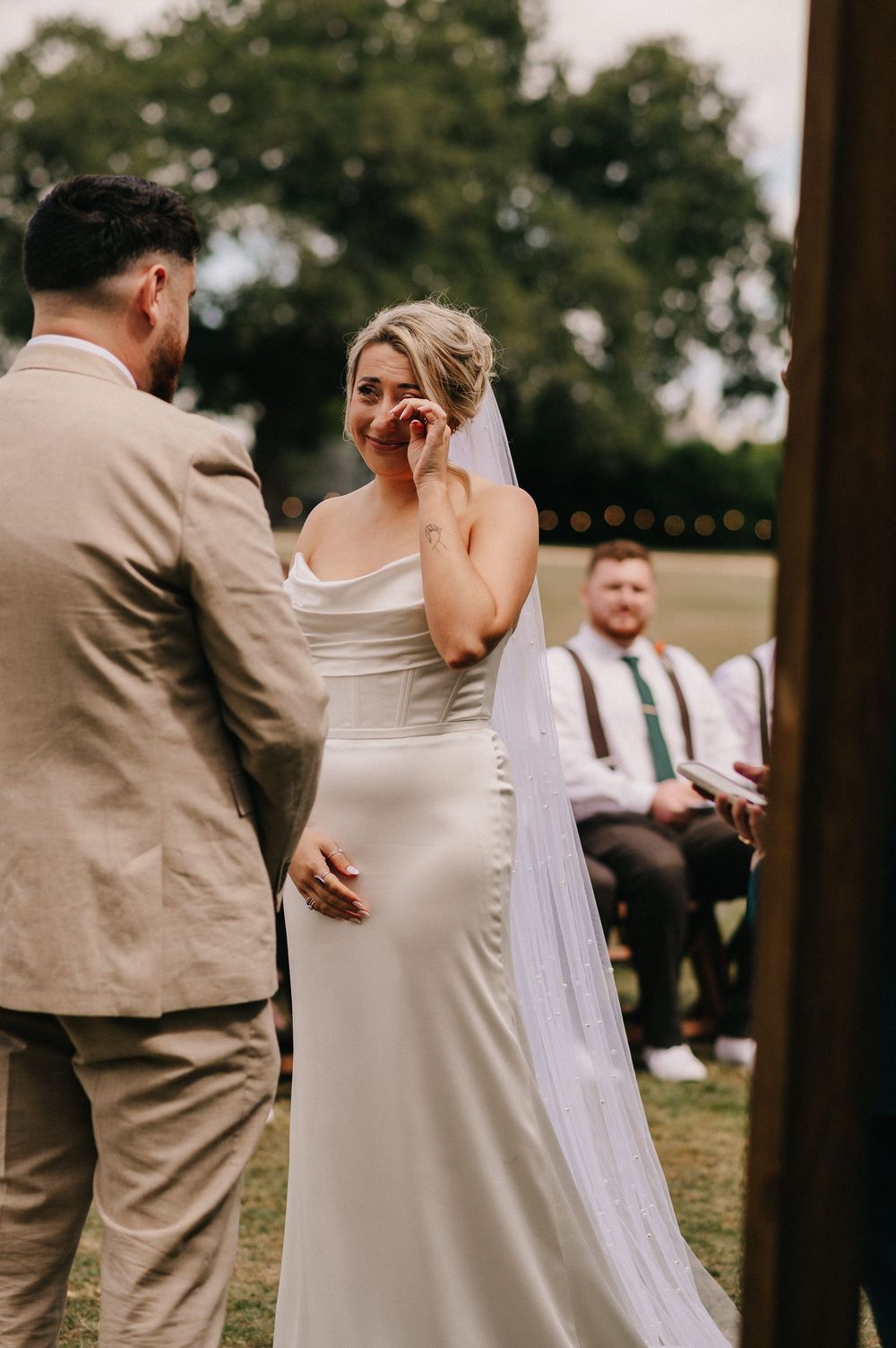 Wedding ceremony guests gather under trees at outdoor venue with wooden chairs and floral arrangements.