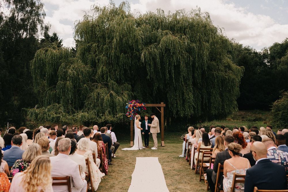 Outdoor wedding ceremony under willow trees with guests seated on white chairs along a path leading to the altar.