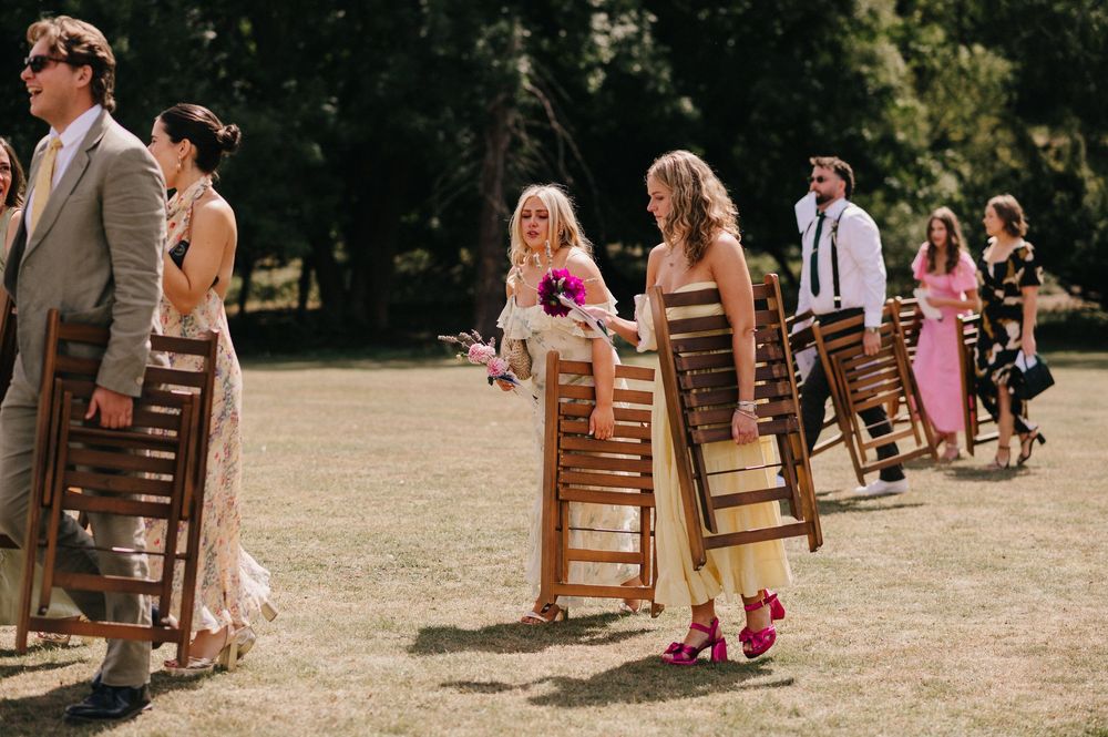 Wooden chairs arranged in rows on grass for an outdoor wedding ceremony with guests gathering in warm sunlight.