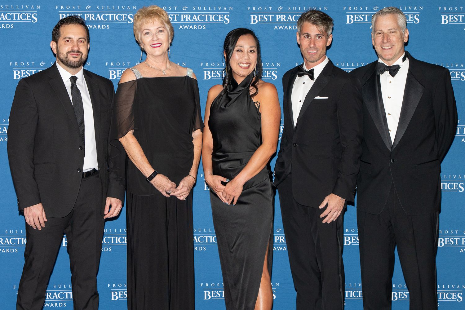 A group of formally dressed people in black suits and evening wear pose together against a blue backdrop at an event.