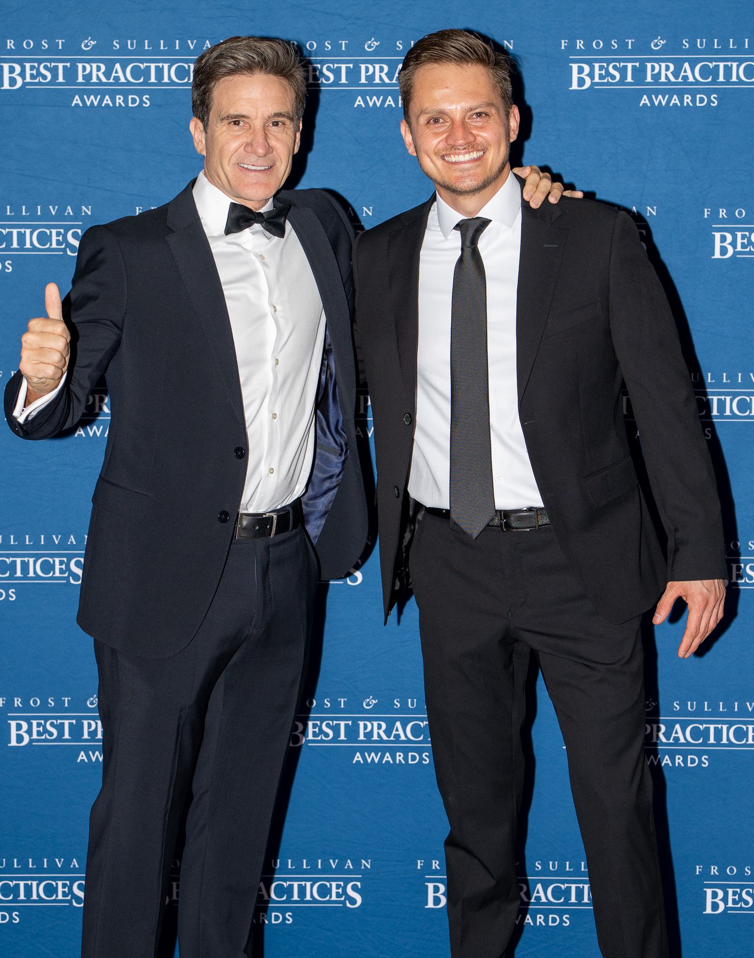 Attendees in formal black suits and ties pose at the Frost & Sullivan Best Practices Awards against a blue backdrop.
