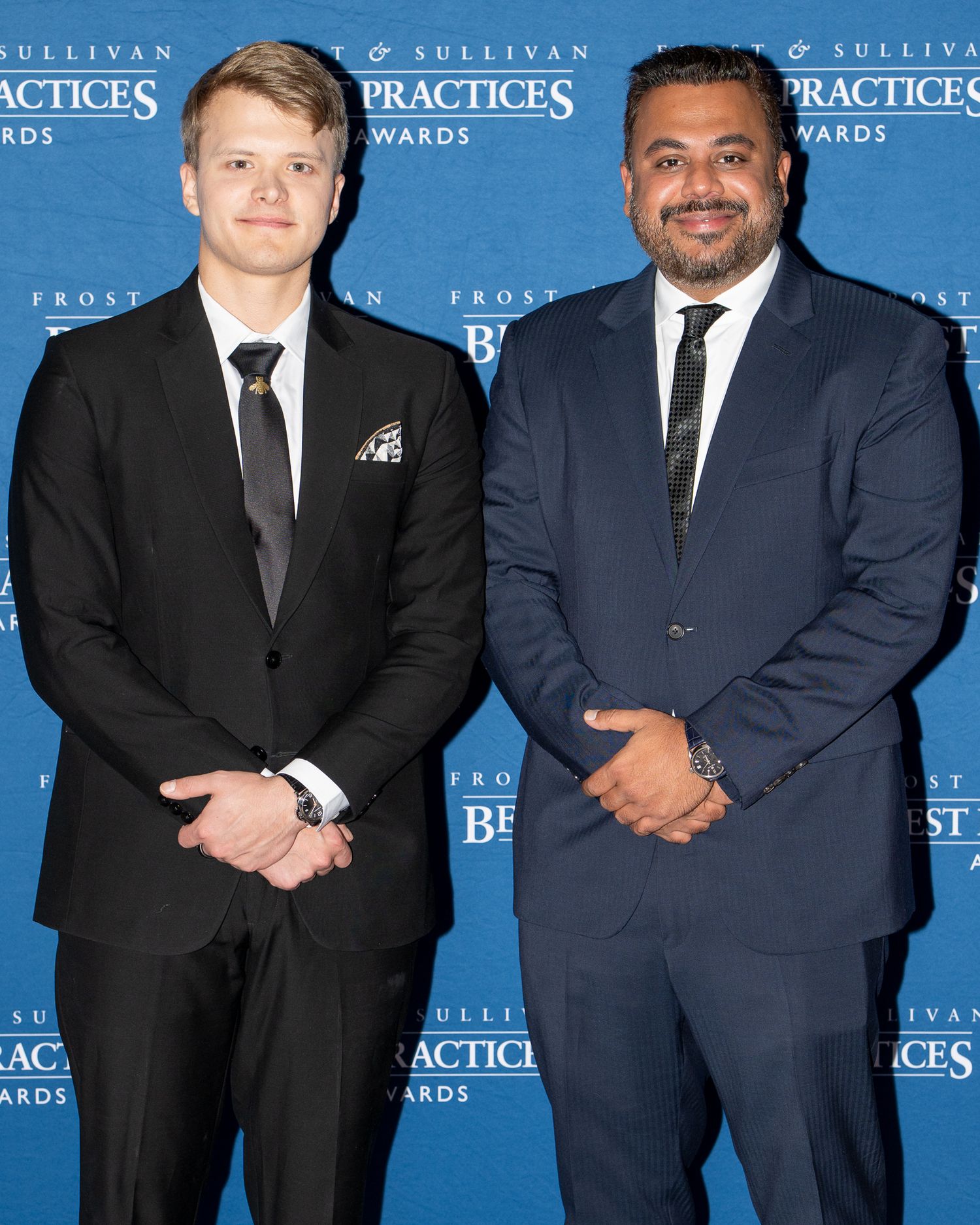 Two business professionals in suits pose together at a Frost & Sullivan Best Practices Awards event with blue backdrop.