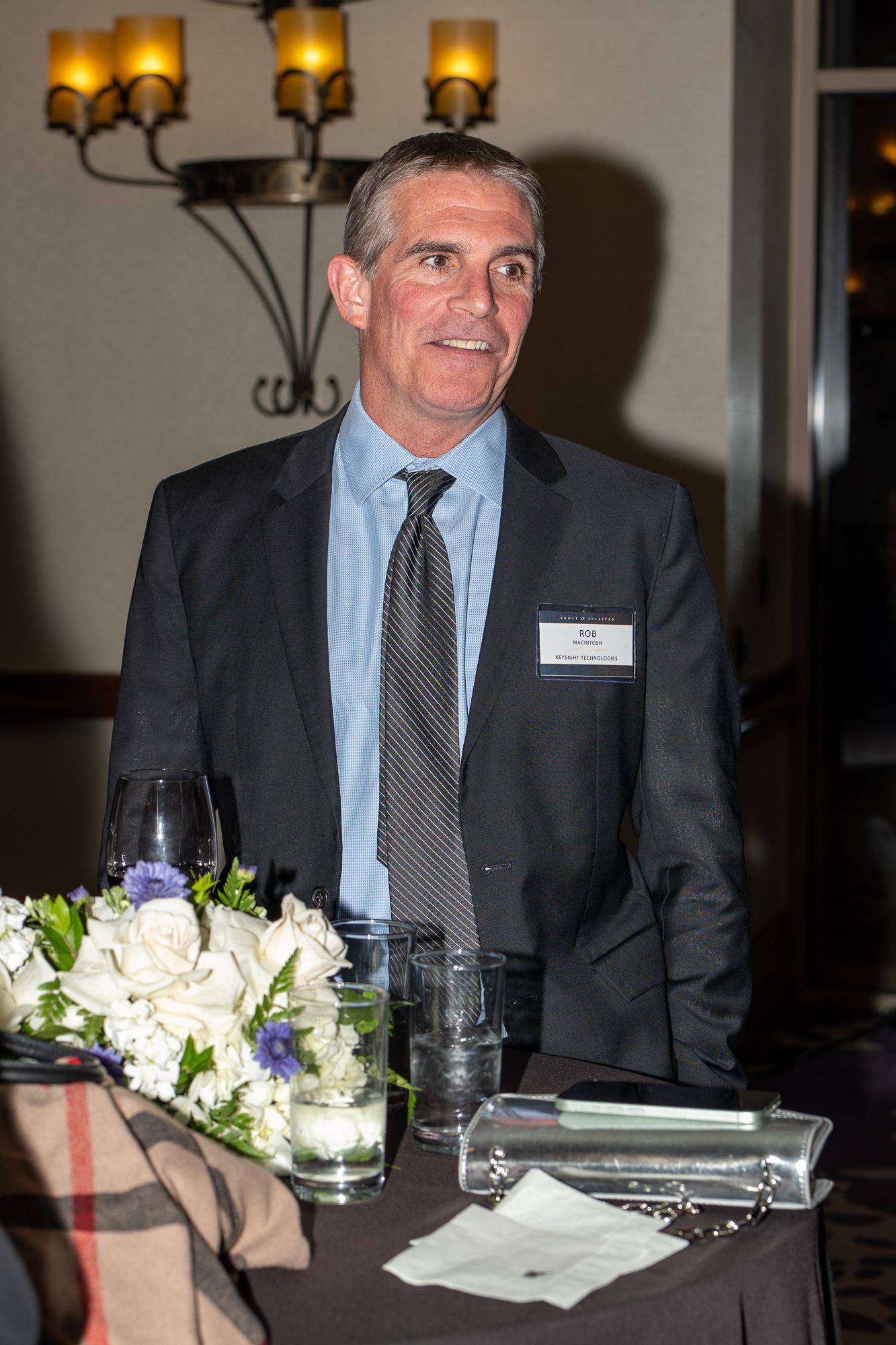 Business executive in dark suit seated at formal dinner table with floral centerpiece and glassware.