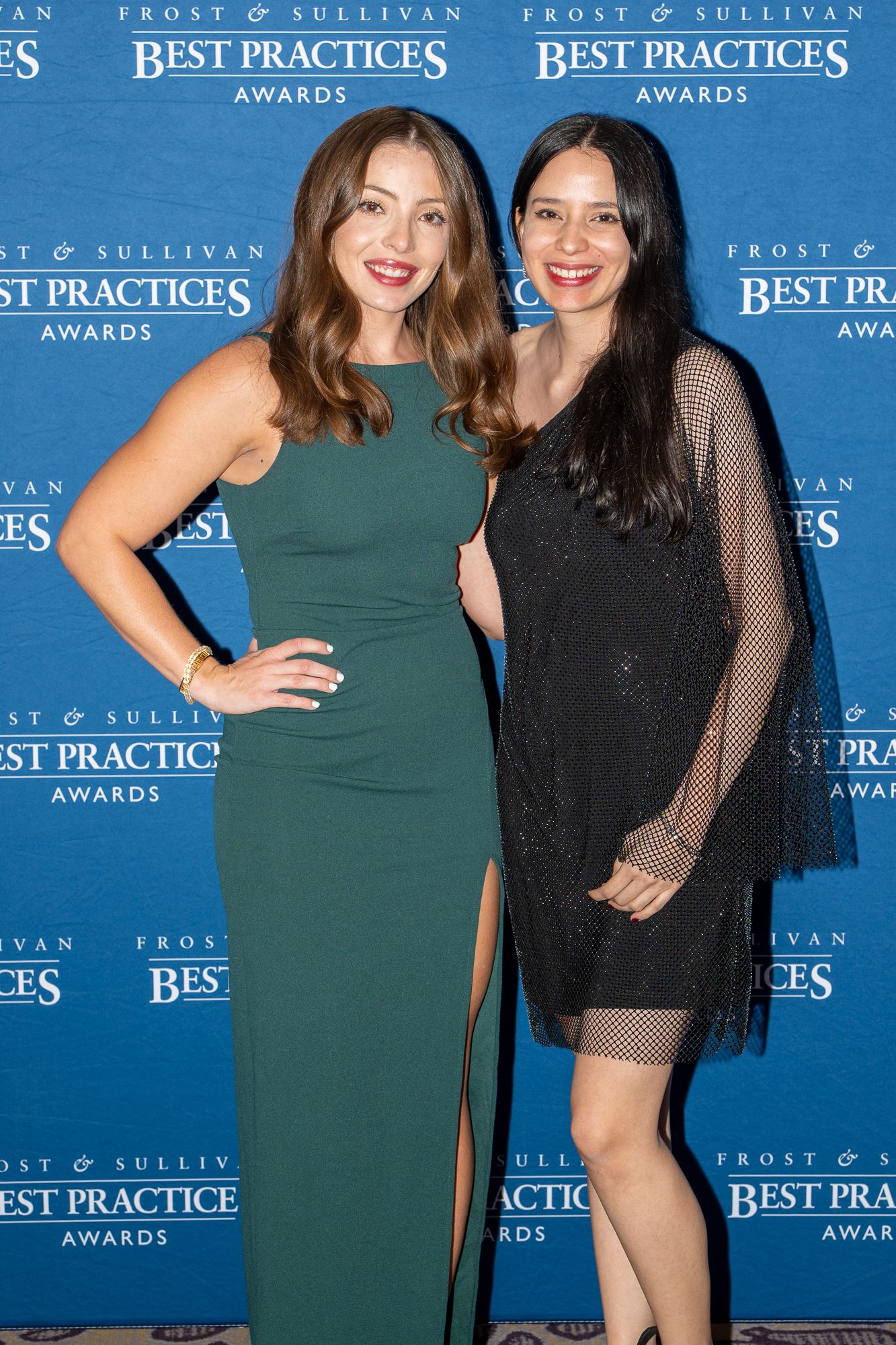Two women in elegant dresses pose together at an awards ceremony with blue backdrop and Best Practices signage.