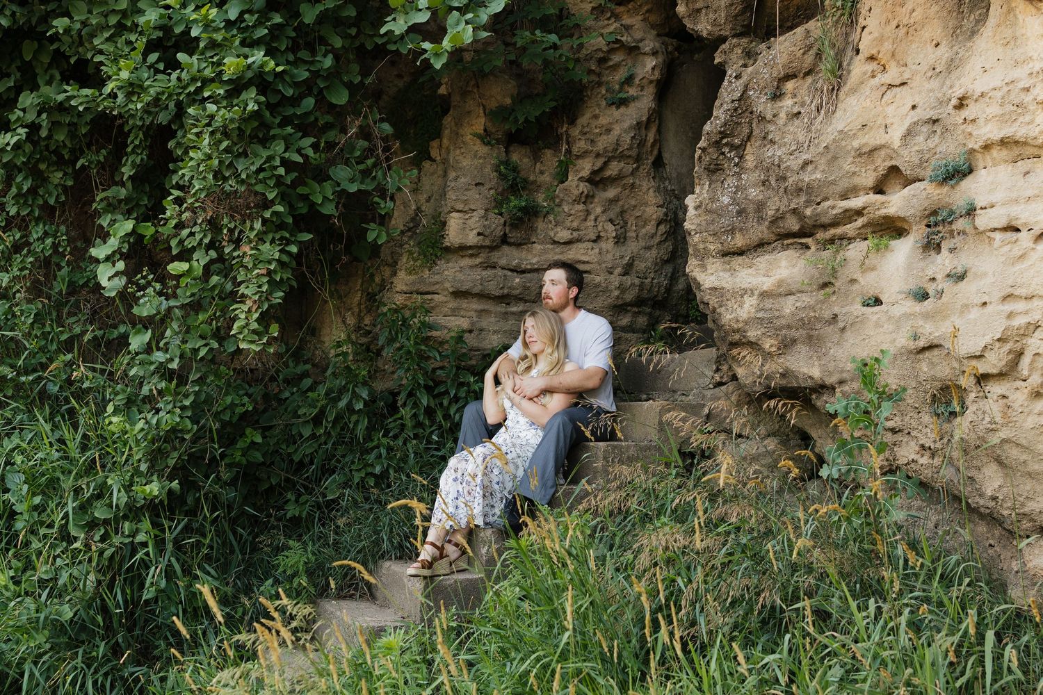 Two people explore the rocky shoreline of a river on a sunny day while examining the natural surroundings.