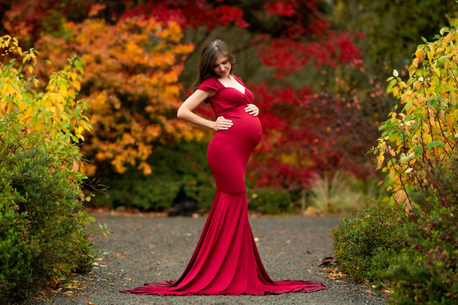 A person in a flowing red gown poses gracefully against vibrant autumn foliage in a garden setting.