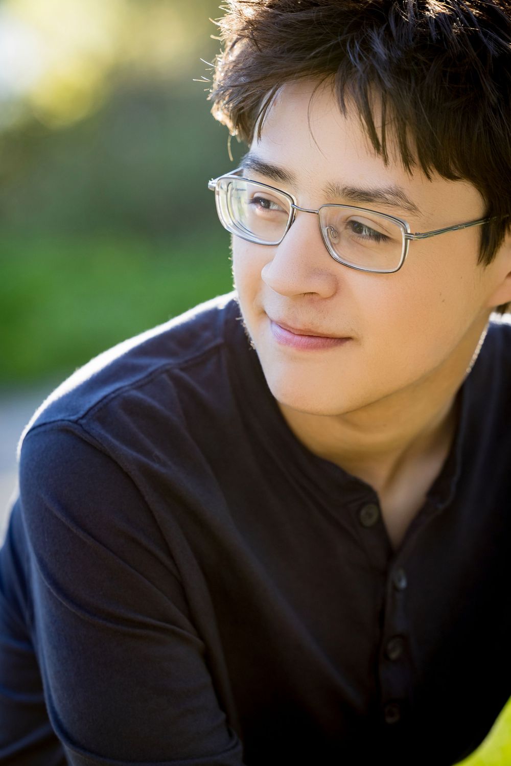 A person wearing glasses and a dark sweater smiles warmly while sitting outdoors in natural lighting.